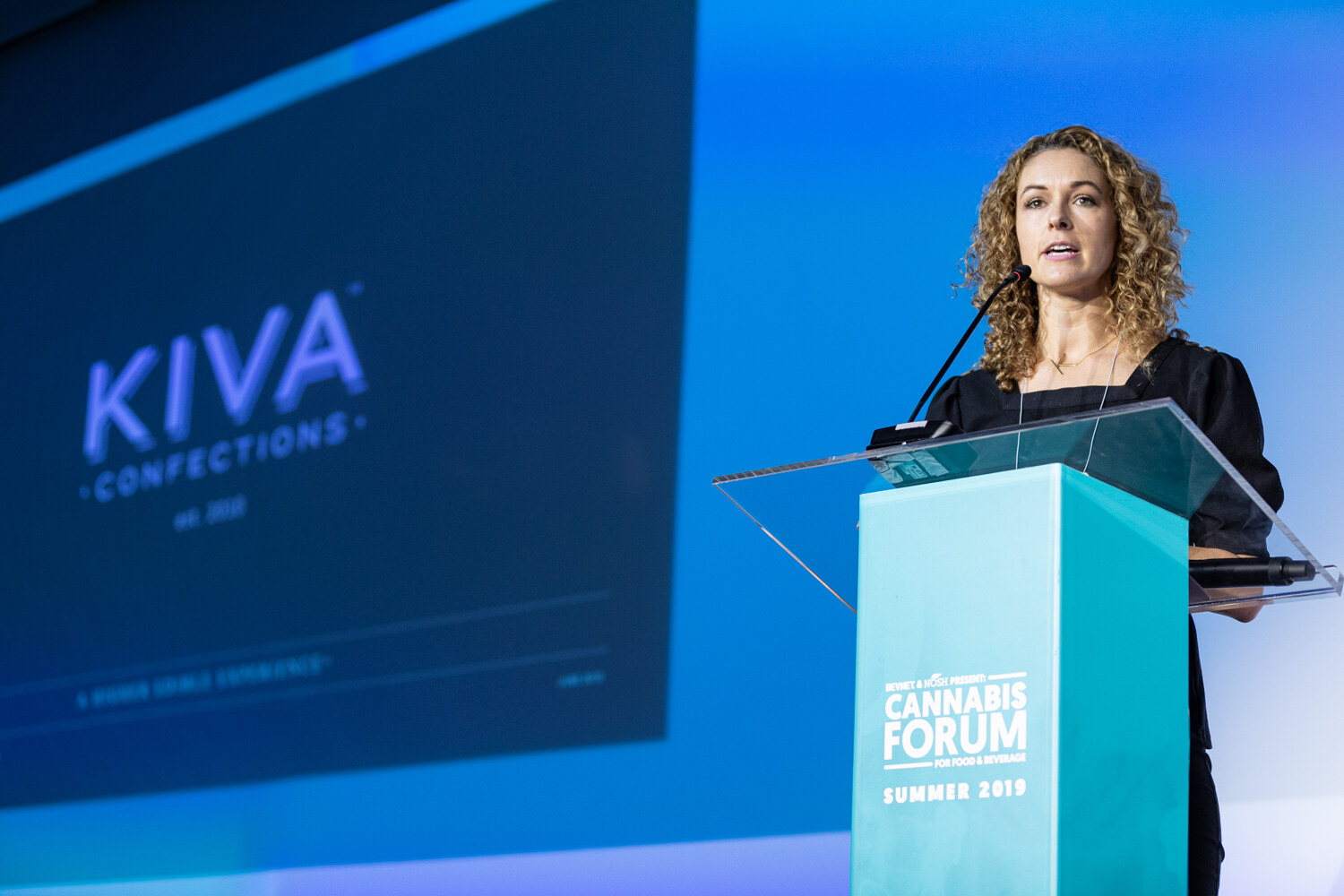 A curly-haired woman in black speaks at a clear podium beside a large dark slide with the KIVA Confections logo.