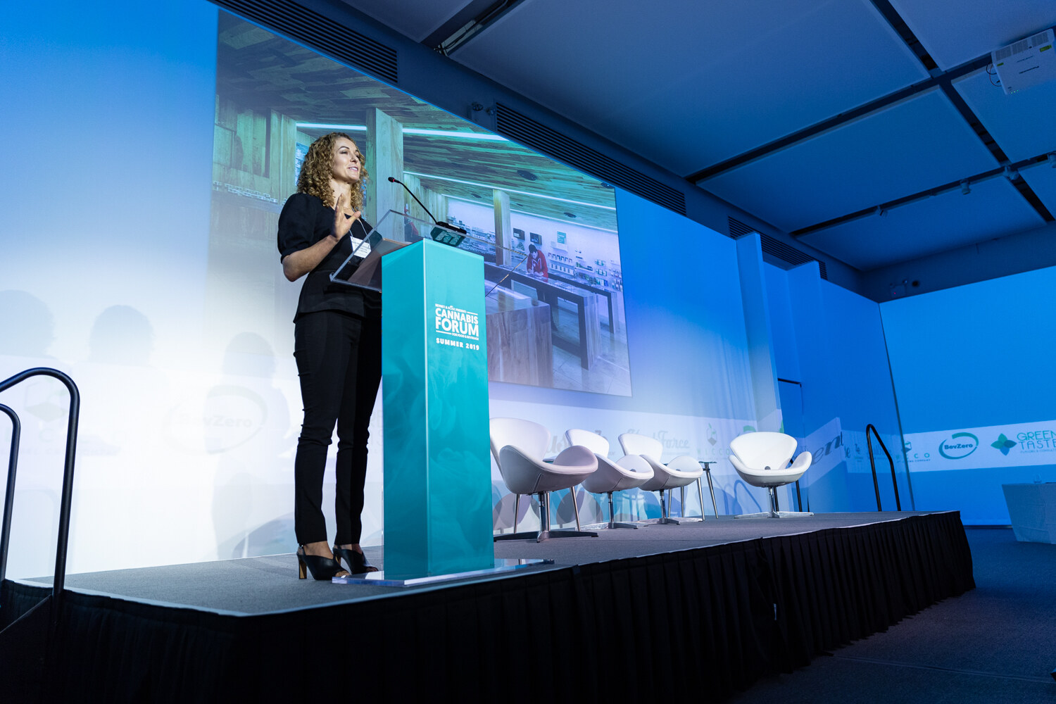A woman in black stands at a teal podium on a stage lit in blue, with a large projected photo of a retail interior behind her and white lounge chairs to the right.