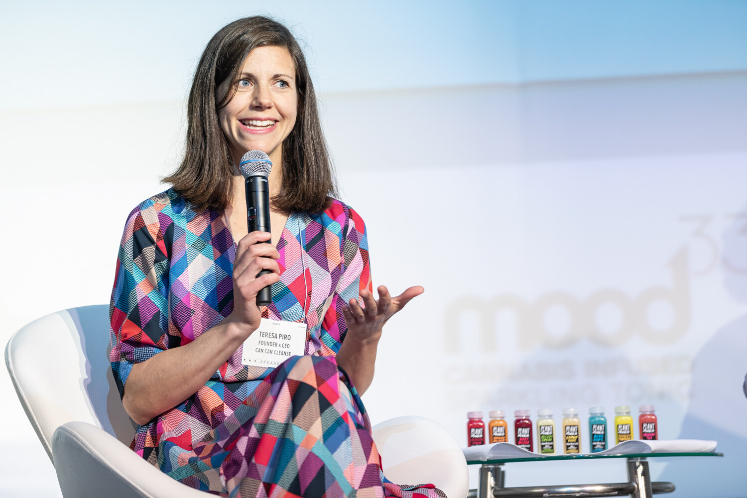 A woman in a colorful geometric-print dress holds a microphone and gestures beside a glass table with a row of small colorful bottles, with a Mood33 backdrop behind.