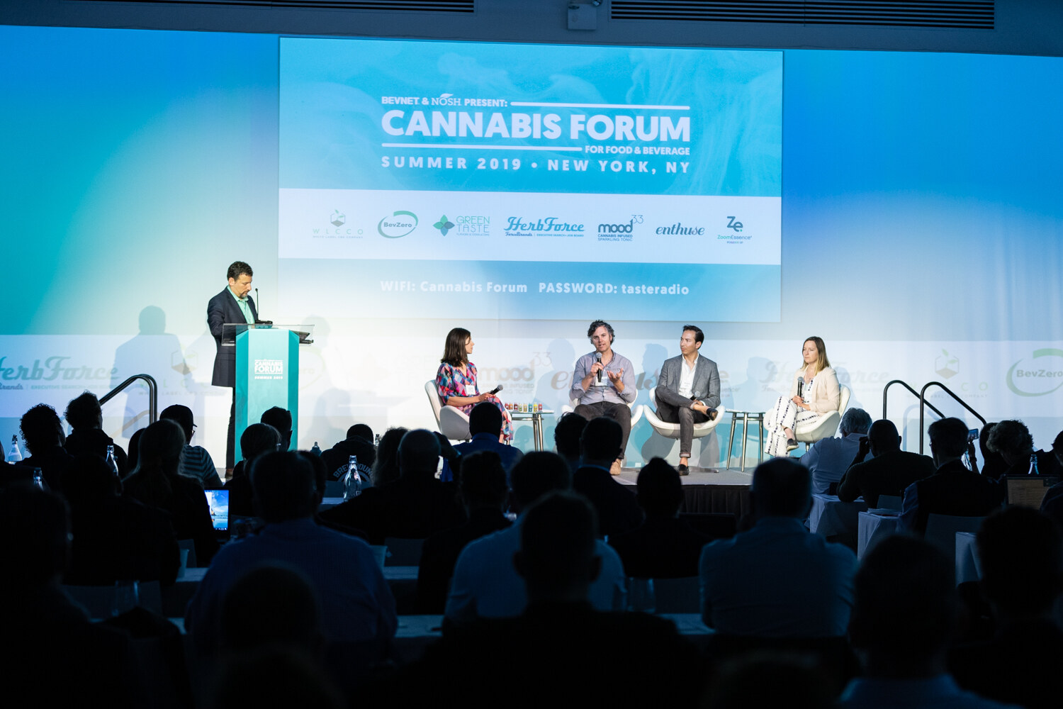 A wide view from the audience shows a host at a podium and four panelists seated on stage under a screen reading Cannabis Forum for Food & Beverage, Summer 2019, New York, NY.