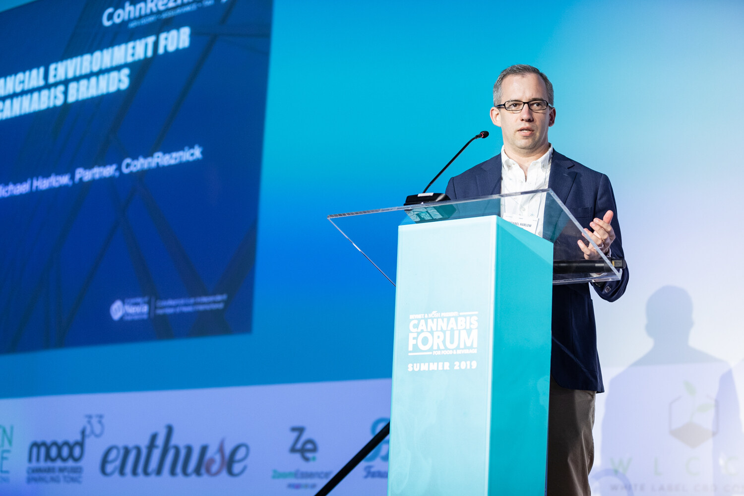 A man in glasses and a navy blazer speaks at a clear podium in front of a blue slide with the CohnReznick logo and the words financial environment for cannabis brands.