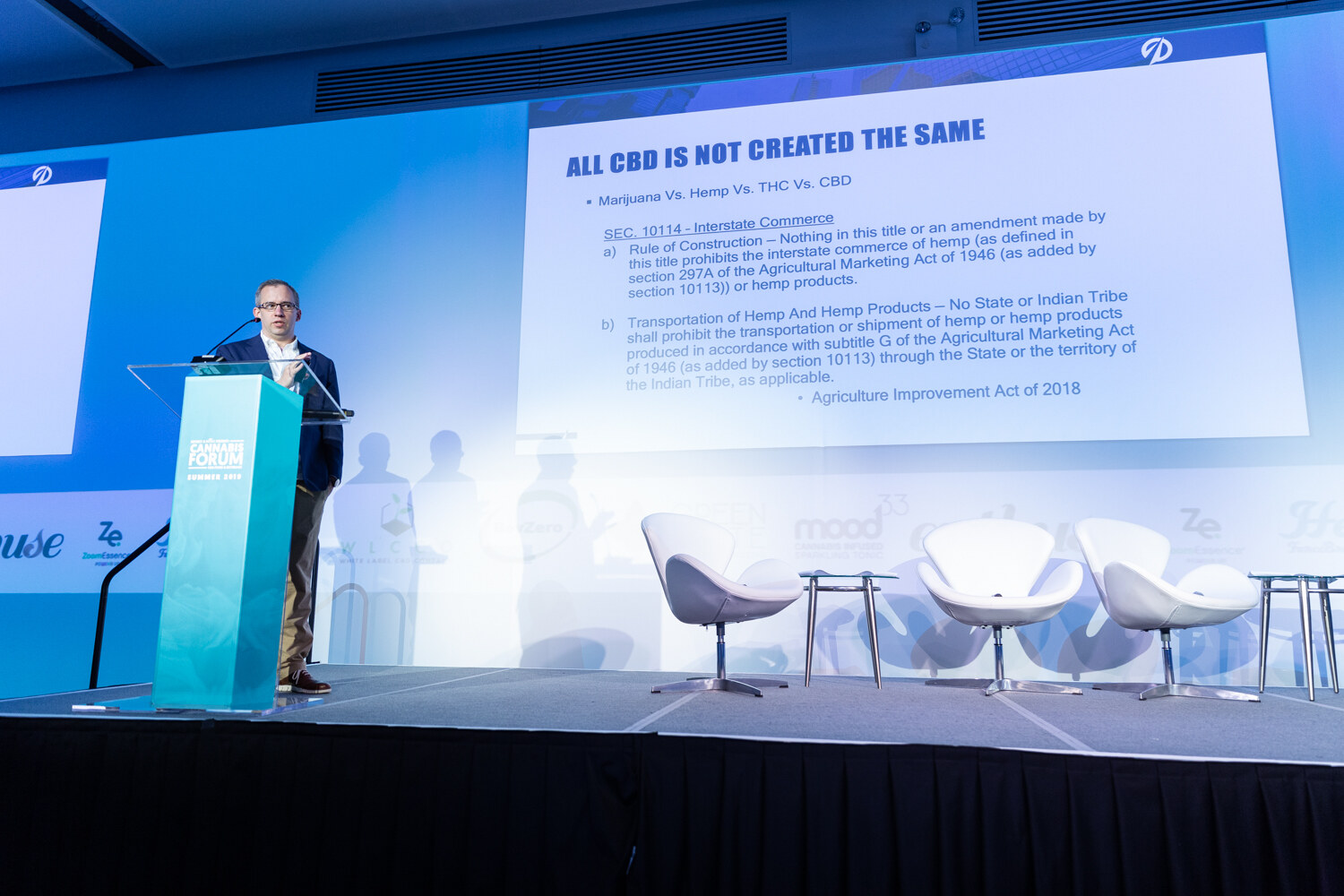 A man in glasses at a teal podium on a long stage beside a large slide headed All CBD Is Not Created The Same, with three white lounge chairs to the right.
