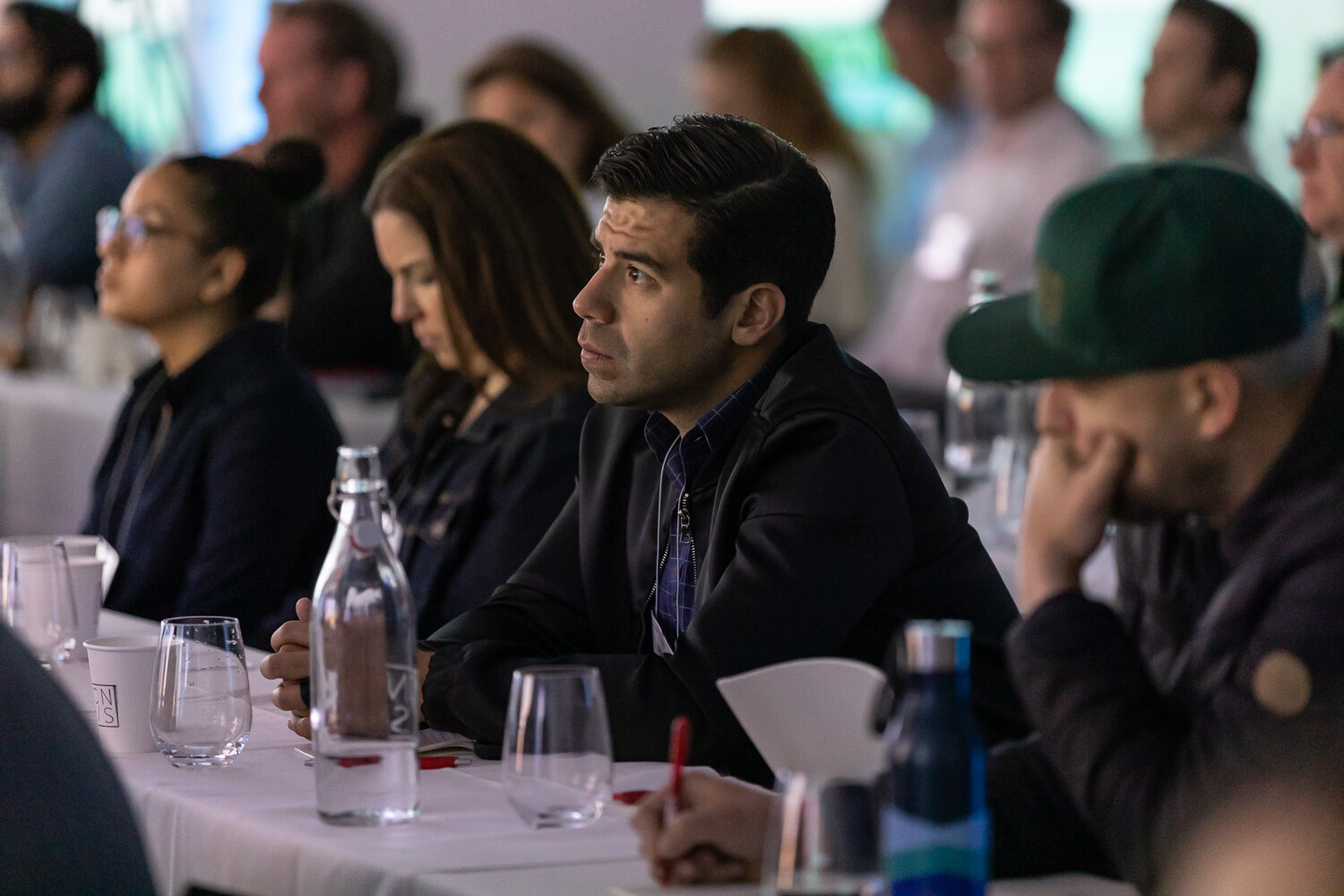 A man in a green cap, a man in a dark jacket and two women sit at a table with water carafes, looking toward the stage.