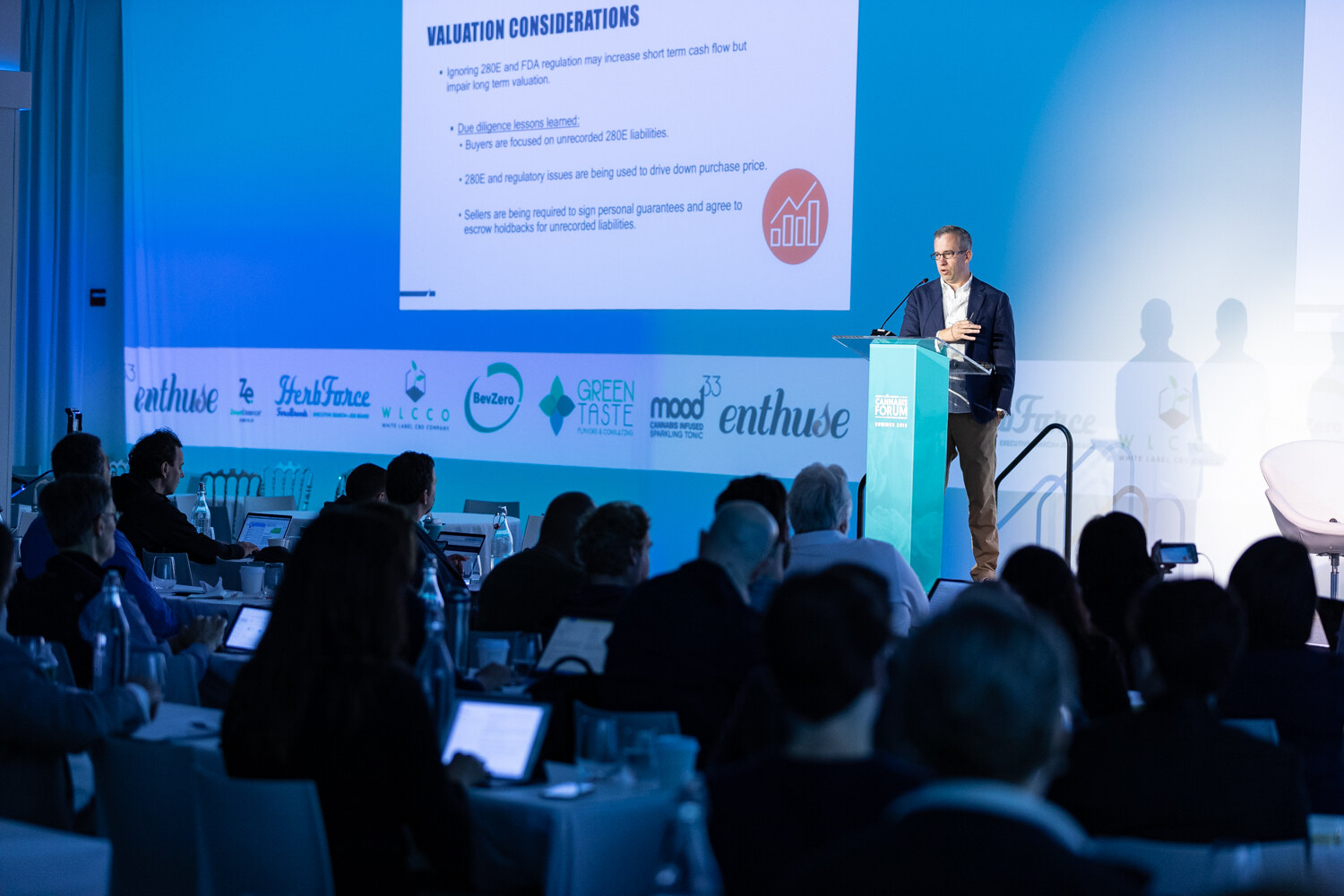 A man at a podium stands beside a slide titled Valuation Considerations, with sponsor logos along the stage and attendees with laptops in the foreground.