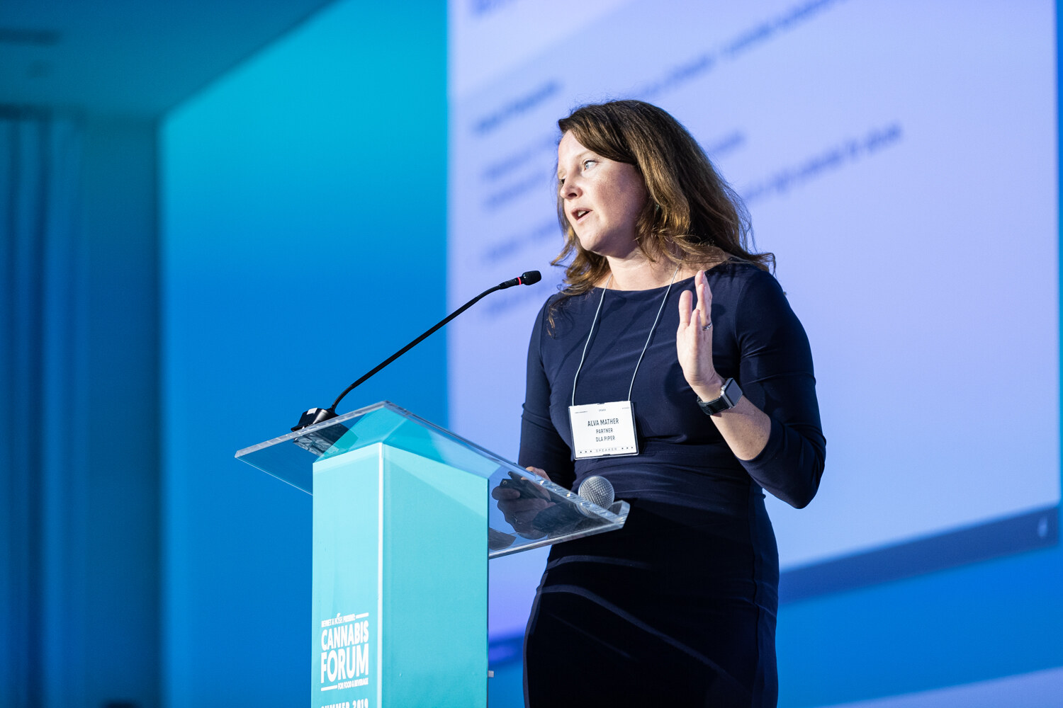 A woman with wavy brown hair and a lanyard speaks into a microphone at a clear podium, with a raised hand and a projected slide behind her.