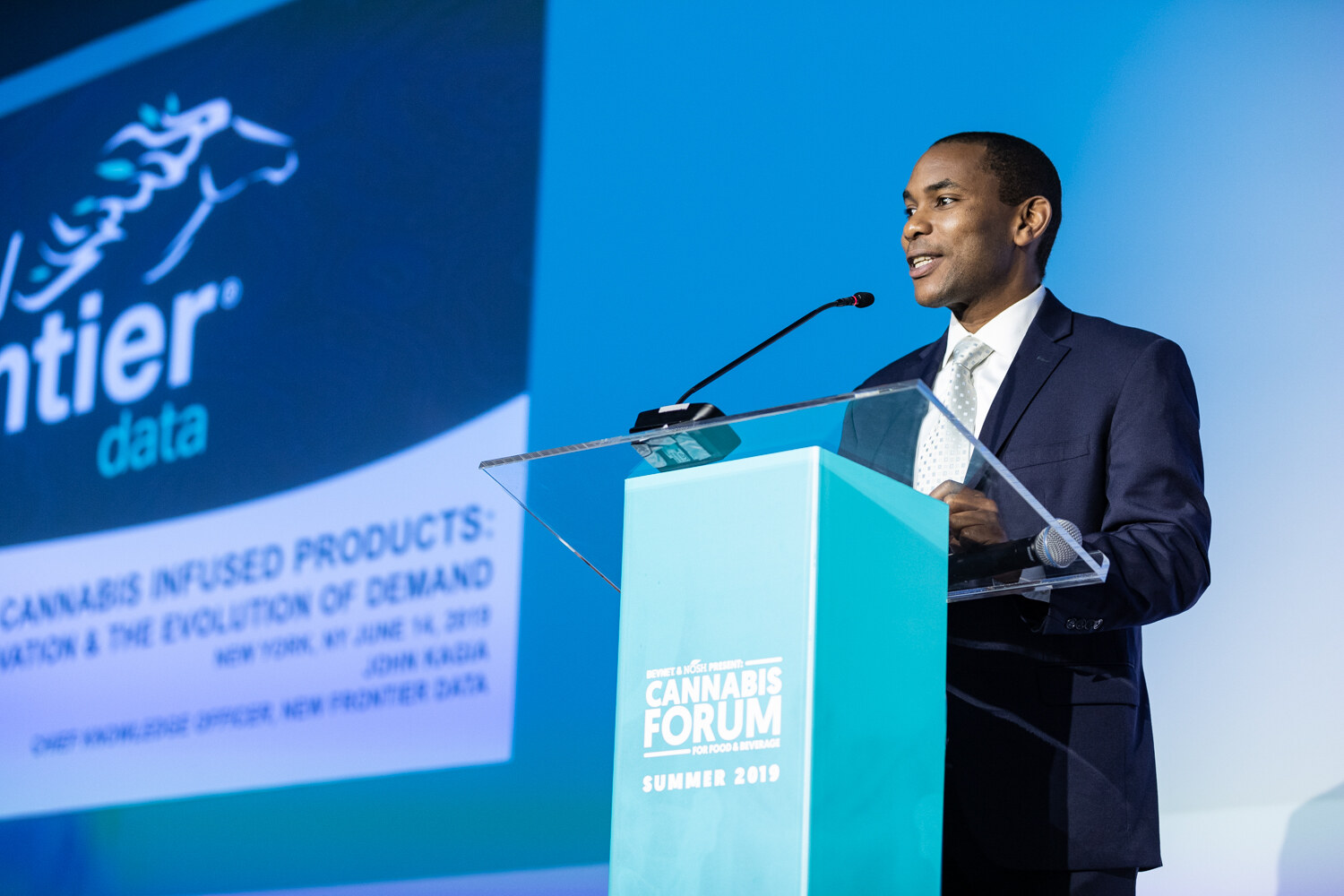 A man in a navy suit speaks at the podium beside a slide with the New Frontier Data logo and the title Cannabis Infused Products: Innovation & the Evolution of Demand, New York, June 14, 2019.