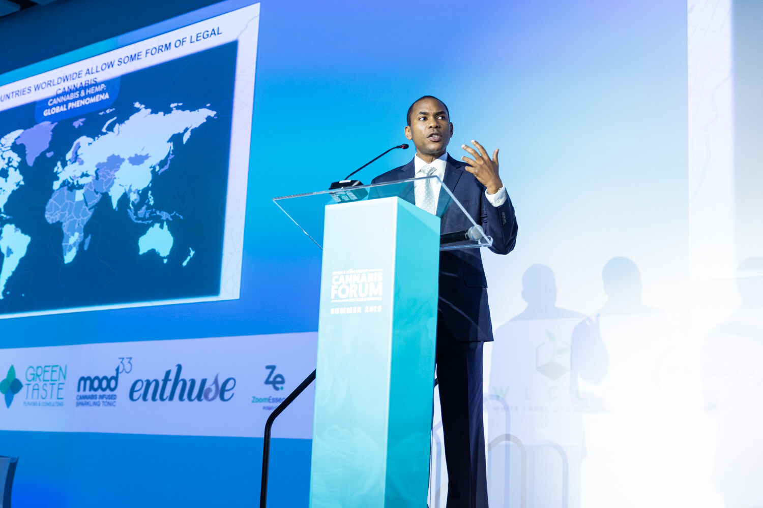 A man in a dark suit and tie speaks at a teal podium next to a large slide with a world map and the heading Countries worldwide allow some form of legal cannabis.