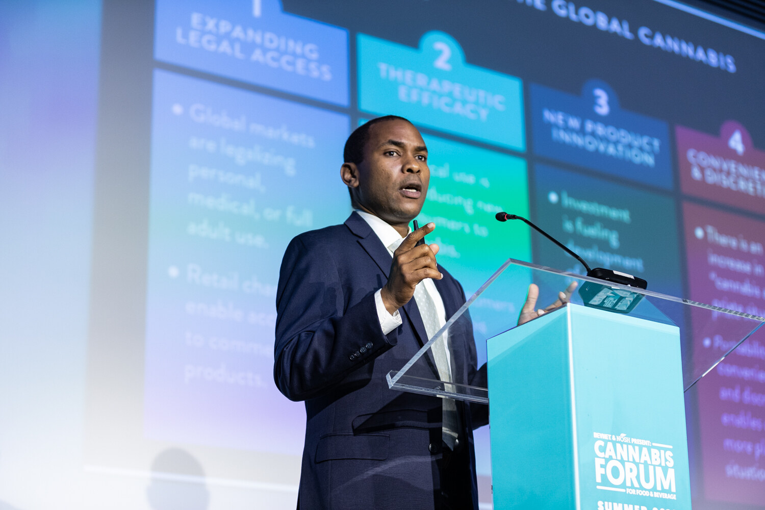 A man in a navy suit gestures near a clear podium in front of a colorful slide listing numbered forces including expanding legal access and therapeutic efficacy.