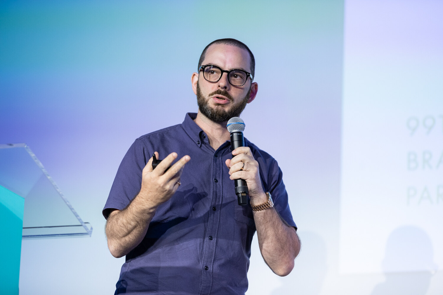 A man with a beard and dark-framed glasses in a navy shirt speaks into a handheld microphone and gestures with his other hand.