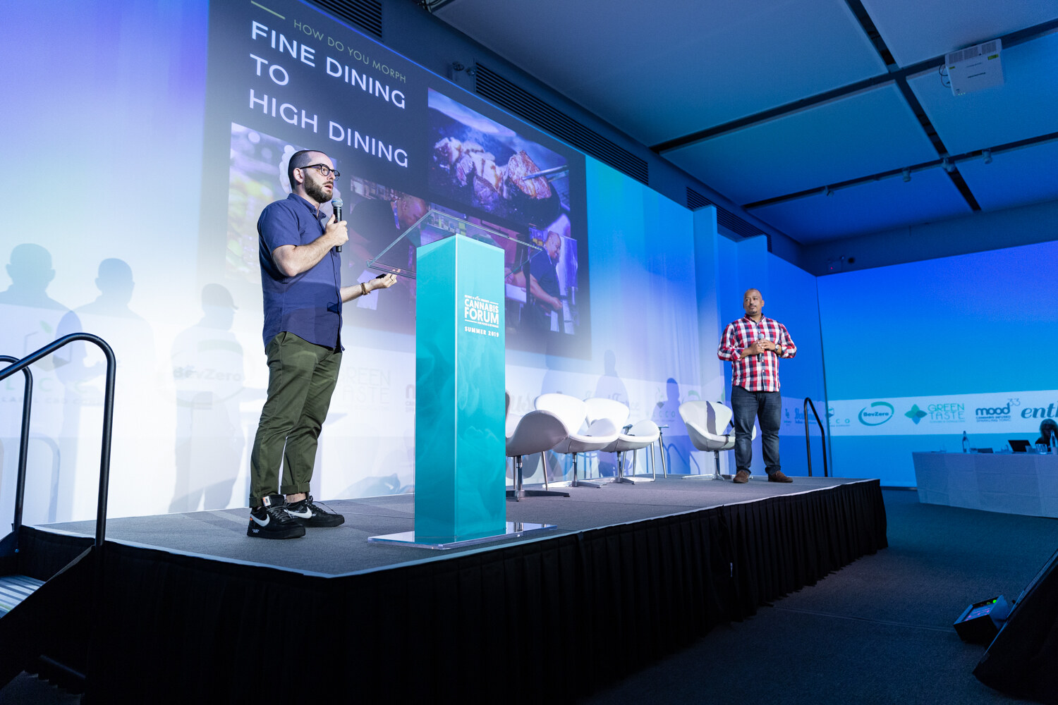 Two men stand on a lit stage, one with a microphone at the podium and one at right in a red plaid shirt, next to a slide reading Fine Dining To High Dining with food photographs.