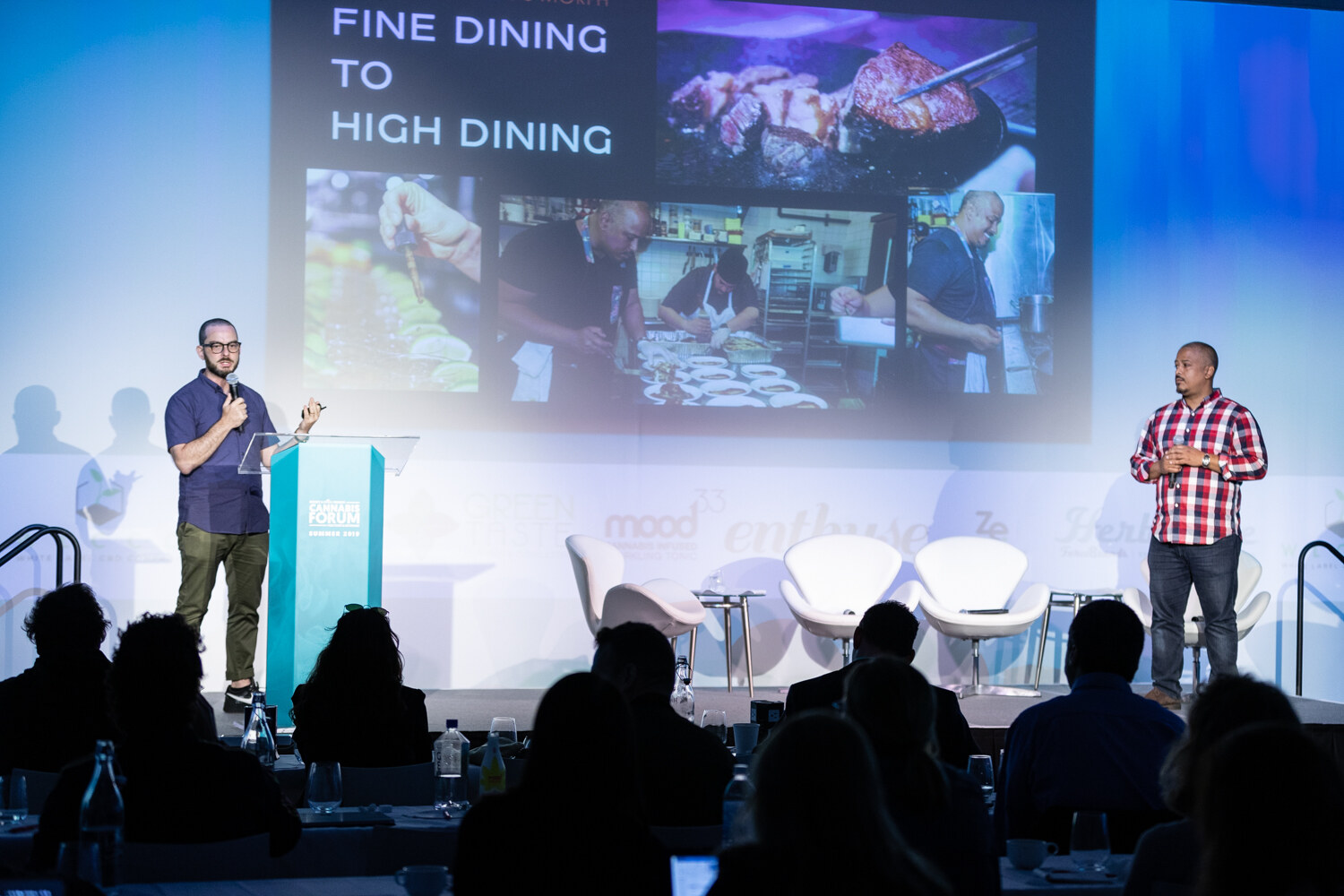Two presenters stand on the stage, one at the podium with a microphone and one in a red plaid shirt at right, below a Fine Dining To High Dining slide, with silhouettes of attendees in front.