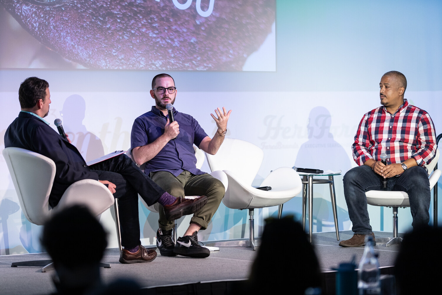 Three men sit on white lounge chairs on stage, a moderator with notes and two presenters, one in a navy shirt gesturing and one in a plaid shirt.