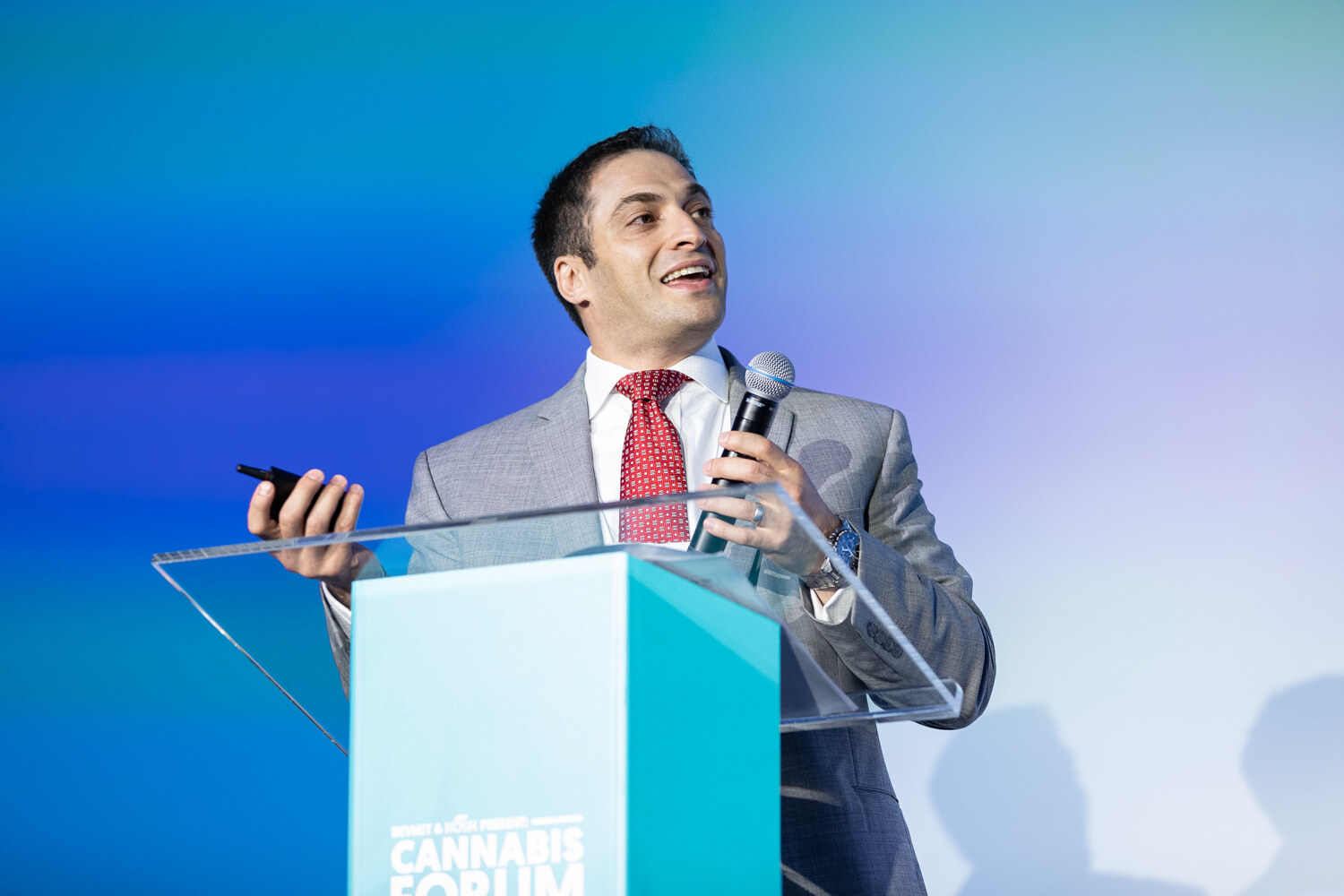 A dark-haired man in a gray suit and red tie holds a microphone and a clicker at a glass podium against a blue backdrop.