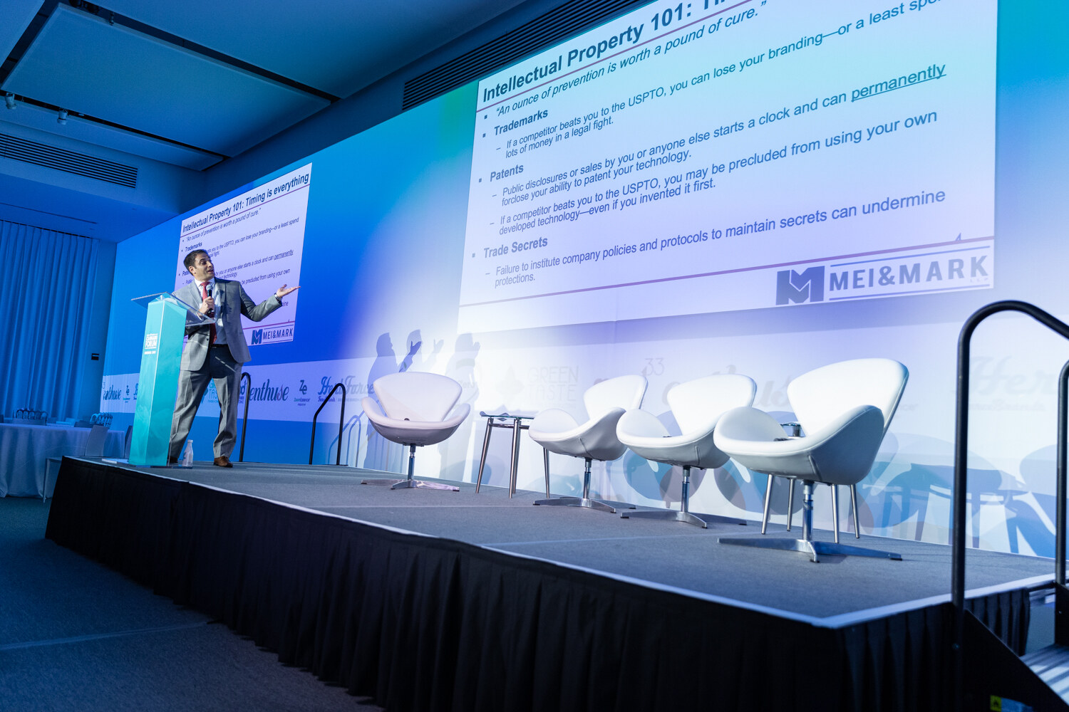 A man at a podium at far left of a long stage beside a large slide headed Intellectual Property 101 with the Mei & Mark logo, and three white lounge chairs on the right.