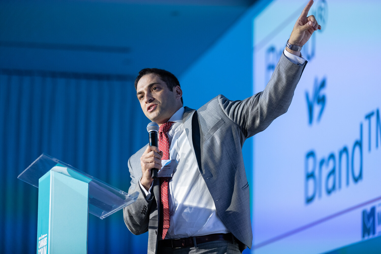 A man in a gray suit and red tie raises one arm toward a large slide with the words vs and Brand, holding a microphone in his other hand.