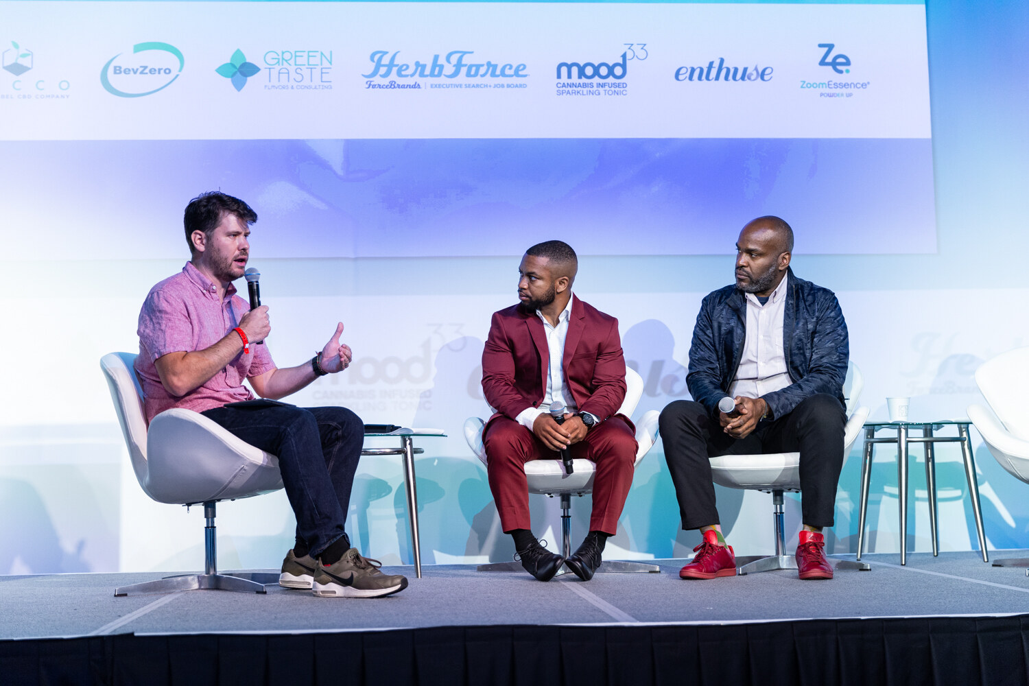Three men sit in white lounge chairs on a bright stage, one in a pink shirt with a microphone and two in burgundy and navy jackets, under a banner of sponsor logos including Green Taste, HerbForce and Mood33.