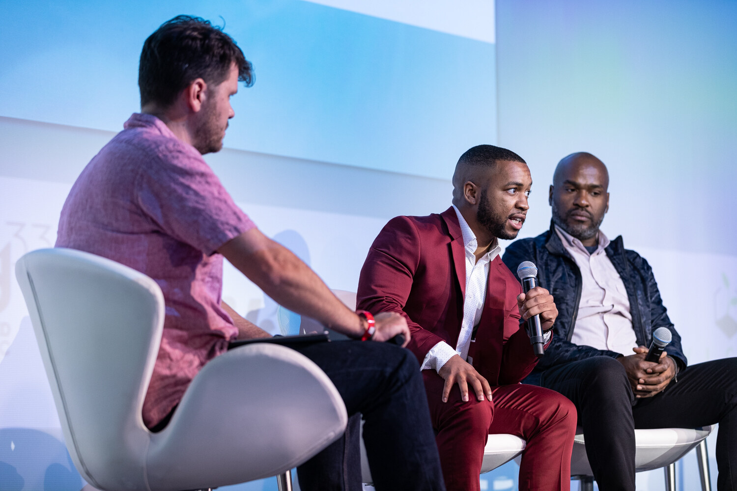 Three men sit in white swan chairs, one in a pink shirt, one in a burgundy suit speaking into a microphone and one in a navy jacket, against a pale blue backdrop.