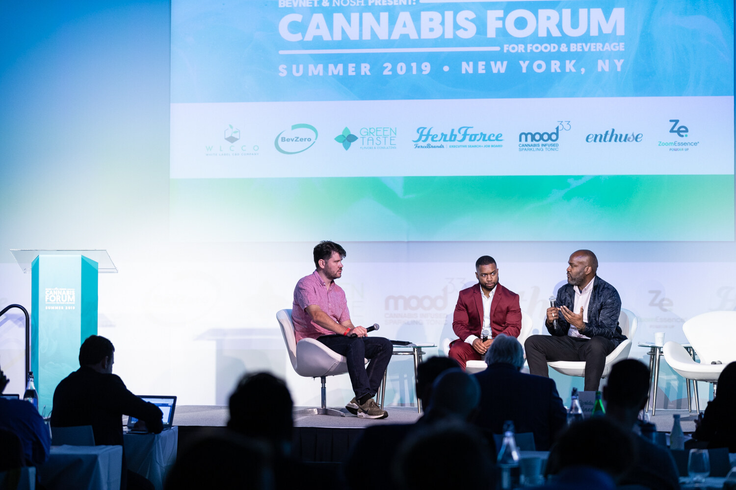 Three men sit on a stage under a screen reading Cannabis Forum for Food & Beverage, Summer 2019, New York, NY with sponsor logos, with dark silhouettes of attendees in front.