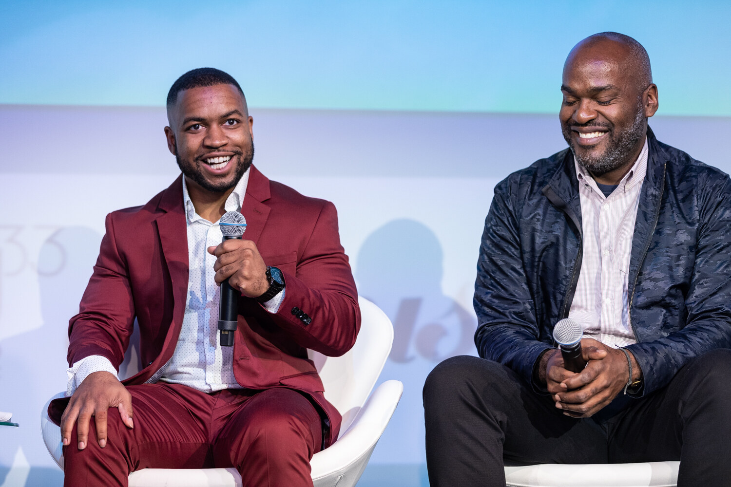 A man in a burgundy blazer smiling with a microphone and a man in a navy jacket laughing with his eyes closed, sitting side by side in white chairs.