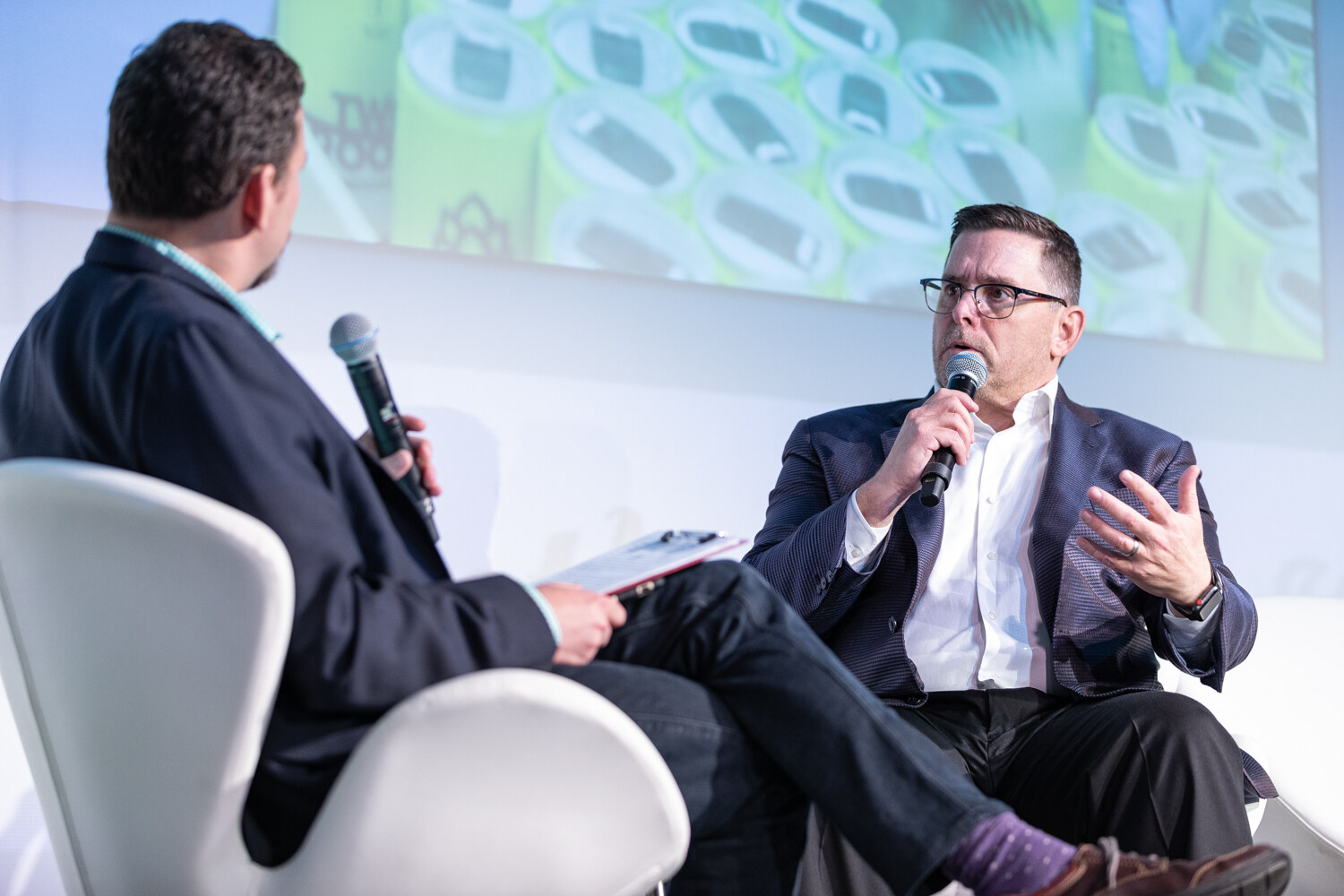 A man in a navy blazer and glasses speaks into a microphone while the moderator, seen from behind, holds notes in a white swan chair, with a screen of green cans above.