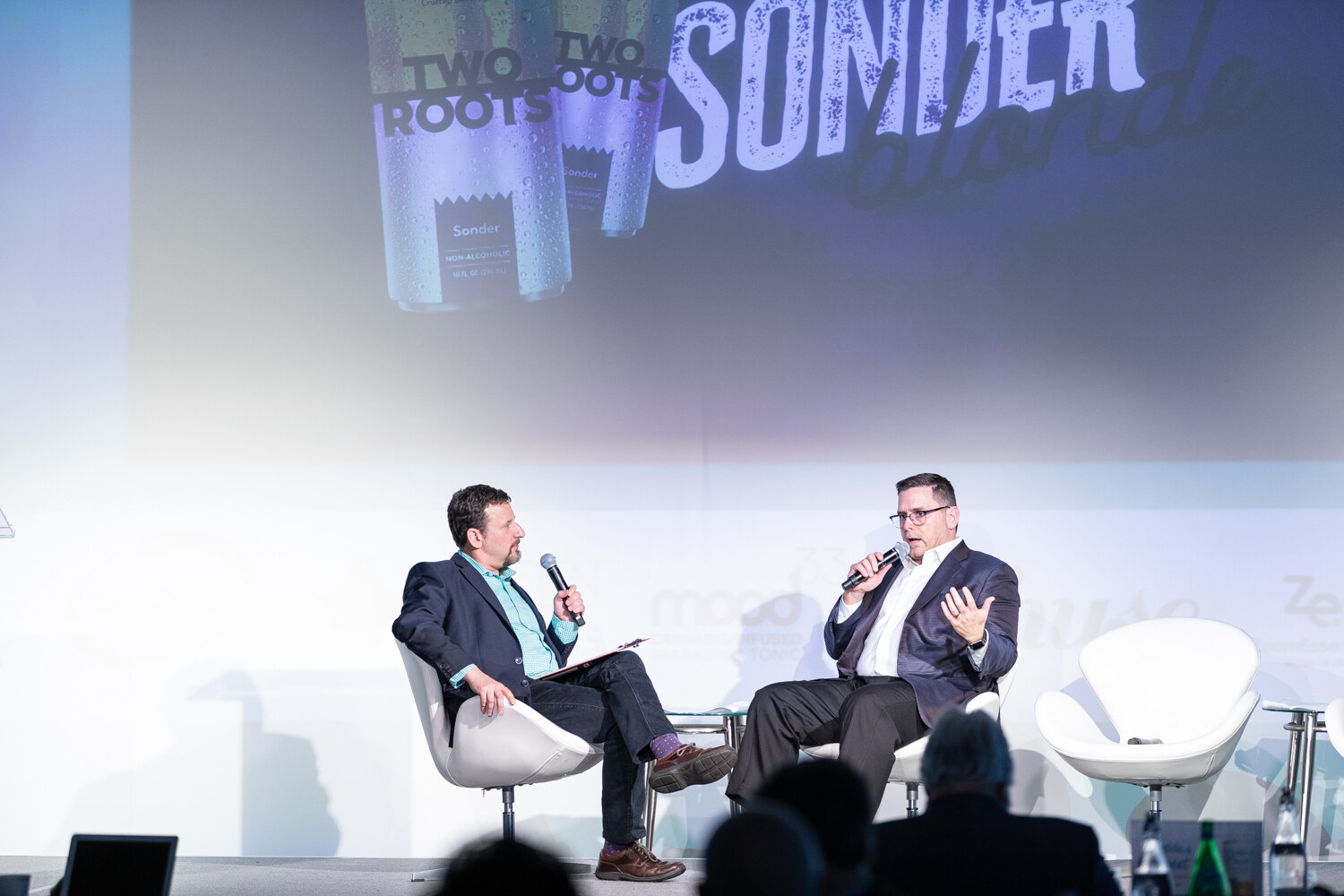 Two men in white lounge chairs hold microphones on a stage below a large screen showing cans labeled Two Roots and the word Sonder.