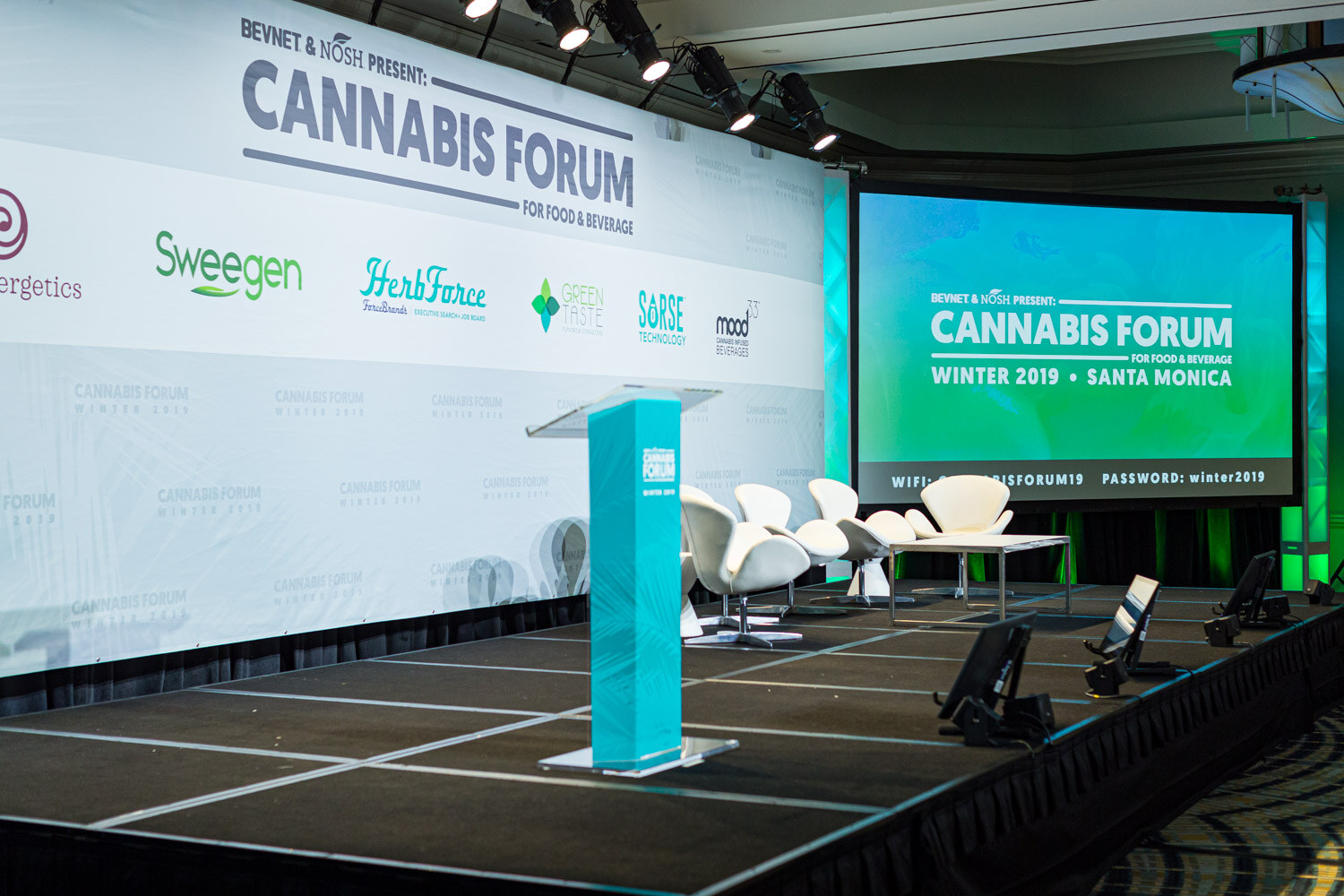 An empty stage with a teal podium, white swan chairs and a screen reading Cannabis Forum for Food & Beverage, Winter 2019, Santa Monica, in front of a sponsor logo backdrop.