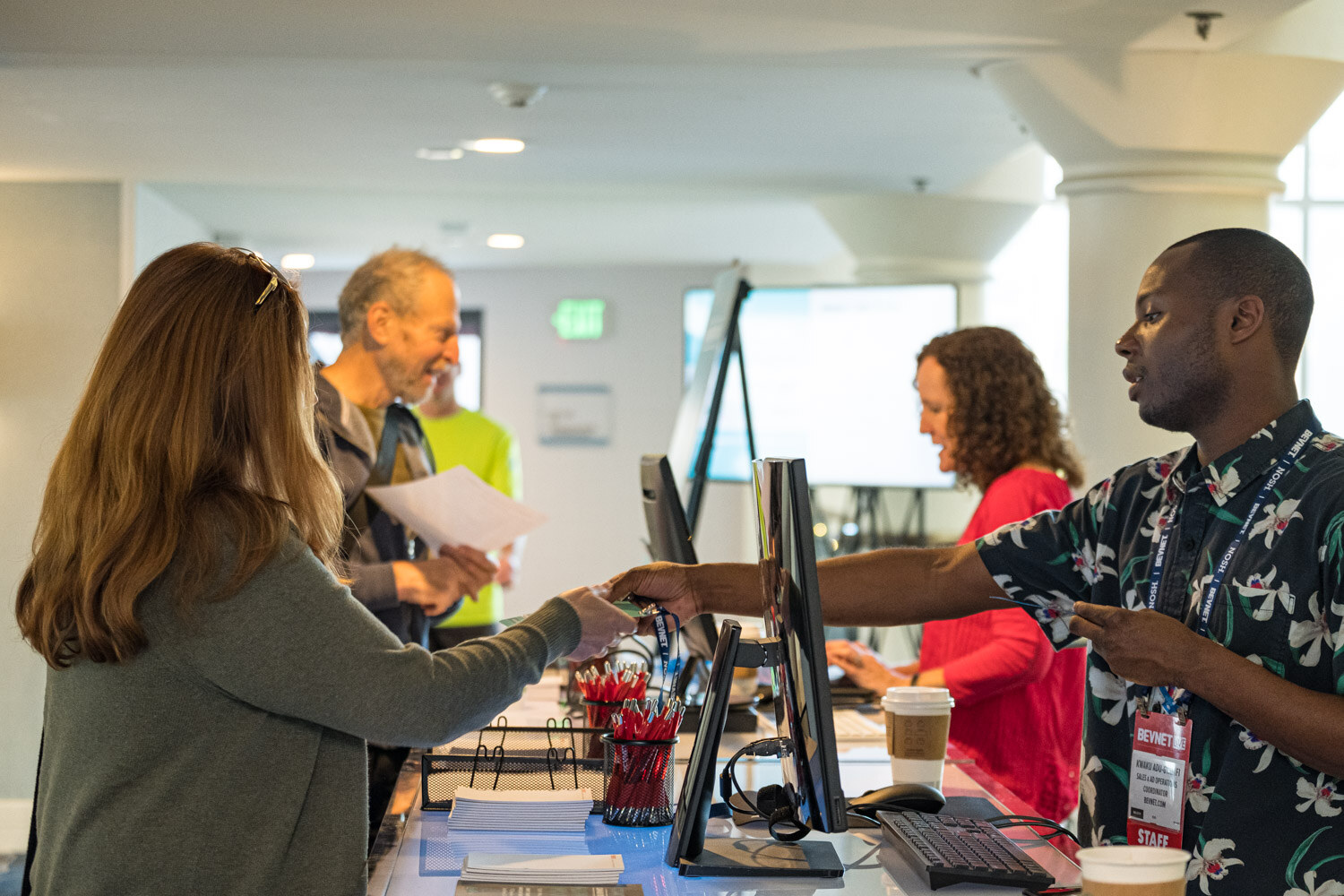 A man in a floral shirt hands a badge across the counter to a woman with long brown hair, with other attendees and staff at the desk behind them in a bright lobby.