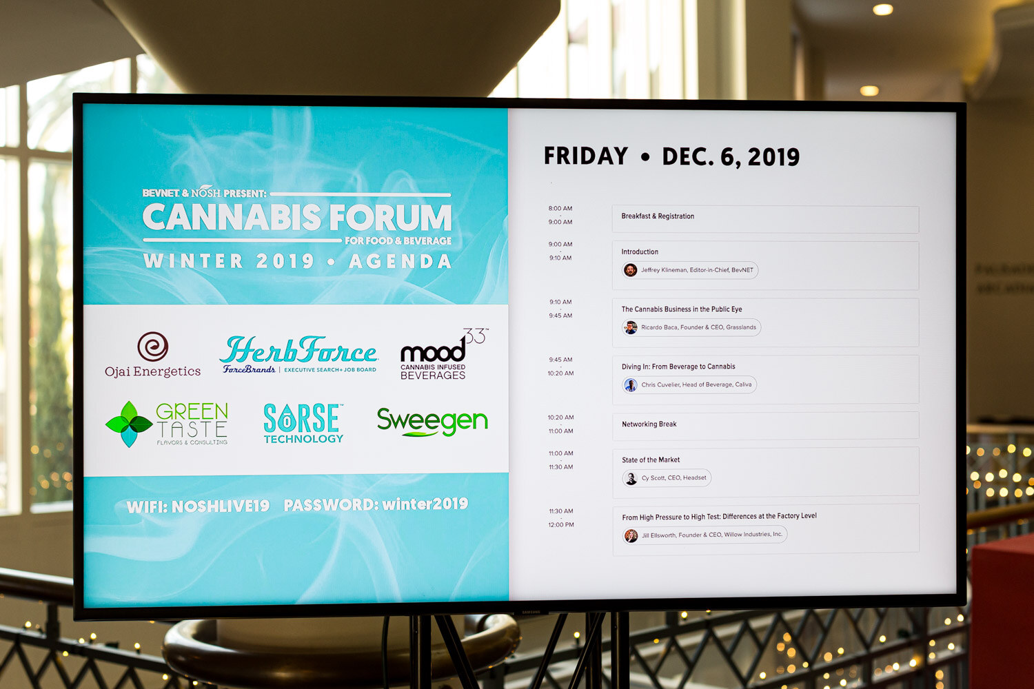 A TV on a stand displays the Cannabis Forum Winter 2019 agenda for Friday, Dec. 6, 2019 with logos for Ojai Energetics, HerbForce, Mood33, Green Taste, Sorse Technology and Sweegen.