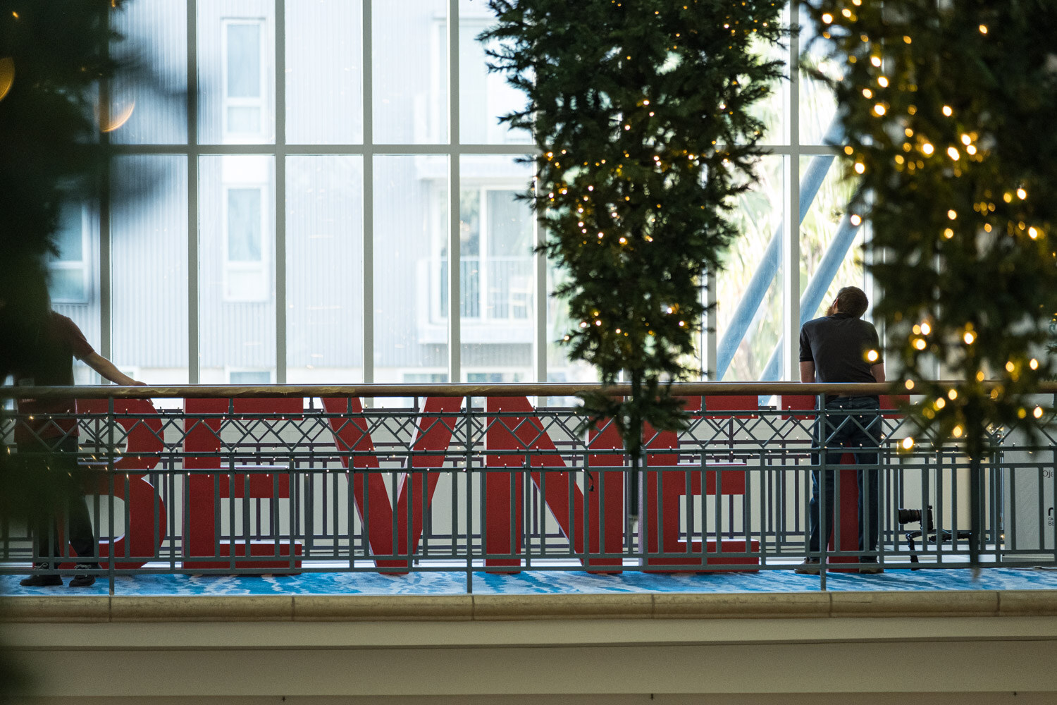 Red block letters spelling BEVNET sit on a metal balcony railing between pine garlands and string lights, with a person standing beyond the railing.