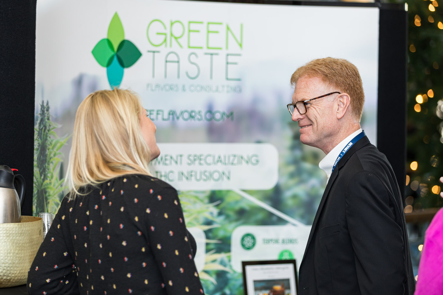 A blond woman and a man in glasses and a dark jacket talk in front of a Green Taste Flavors & Consulting banner.