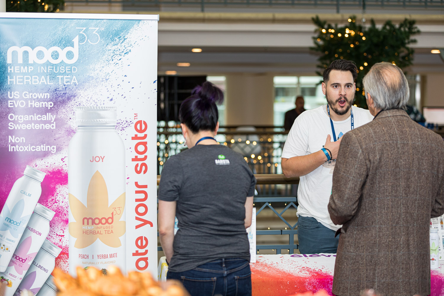 A man in a white t-shirt speaks with two attendees beside a tall Mood33 Hemp Infused Herbal Tea banner showing Joy, Calm and Passion bottles.