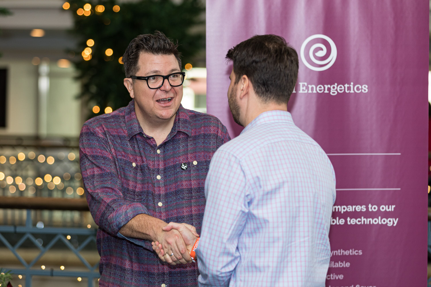 Two men shake hands in front of a purple Ojai Energetics banner in a bright hallway with string lights behind them.