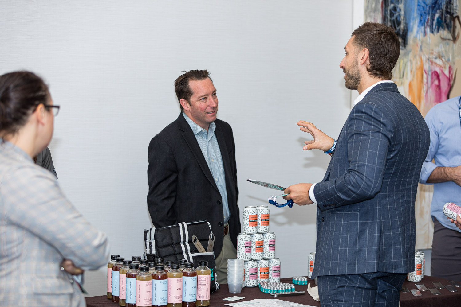 Two men in jackets chat behind a table holding rows of small bottles and cans, with a woman in glasses listening at left.