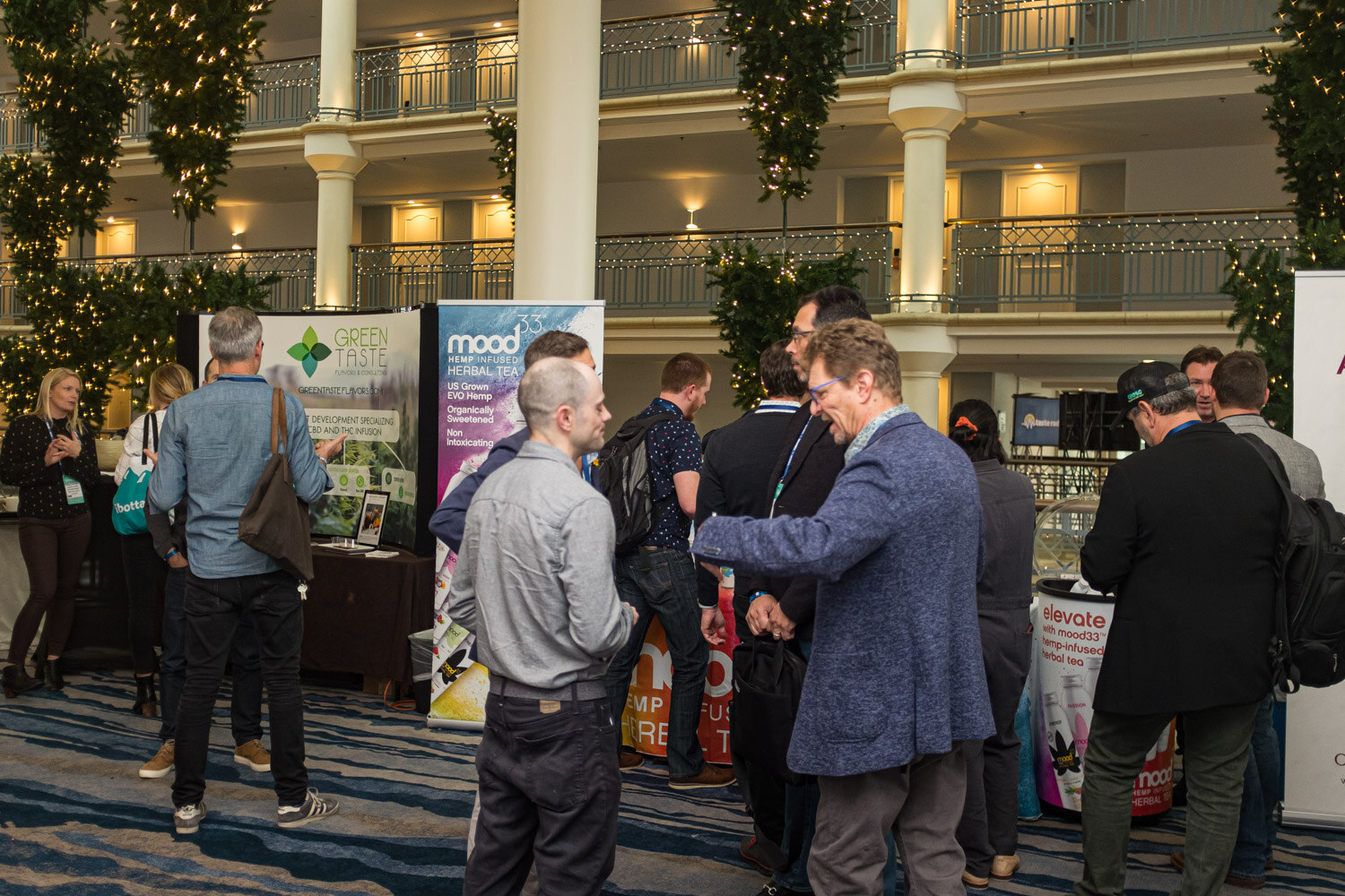 A crowd of attendees talks in front of tabletop displays and pull-up banners including Green Taste and Mood33, with a balcony hung with lights above.