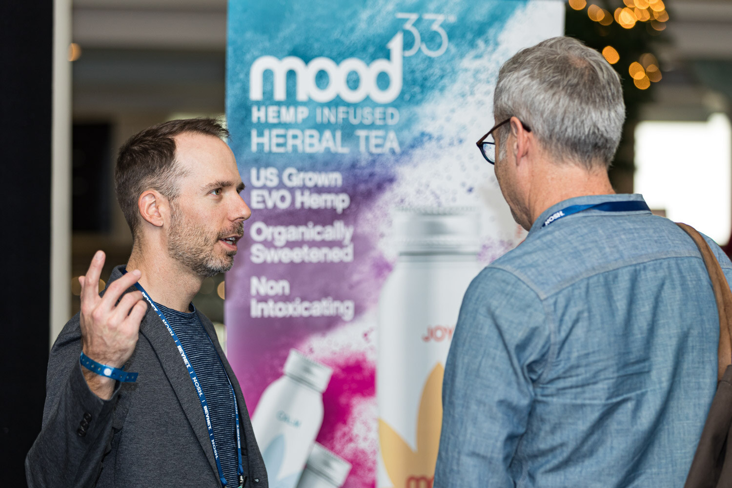 A man with a lanyard gestures as he talks to an older man in a denim shirt, in front of a Mood33 Hemp Infused Herbal Tea banner.