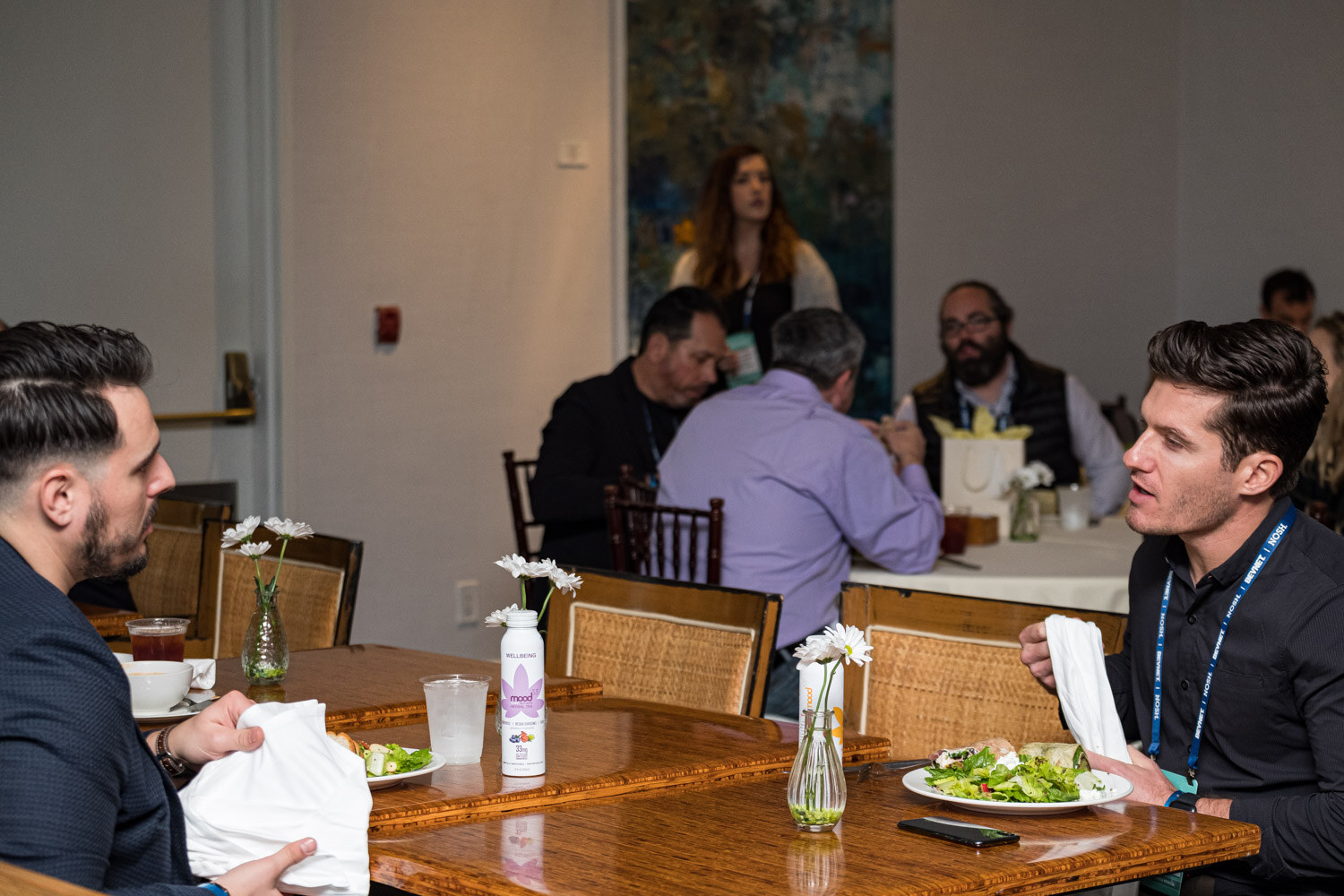 Several attendees in lanyards sit at wooden tables with salads and small flower vases, and a lavender-labeled bottle stands in the foreground.
