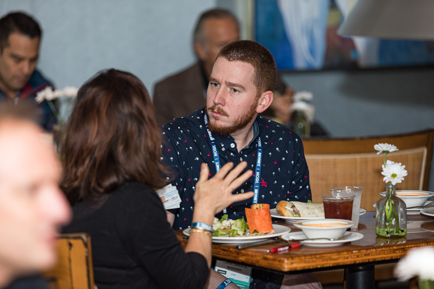 A bearded man in a polka-dot shirt and a lanyard talks with a woman gesturing with her hand across a table of salads and wraps, with another attendee at left.