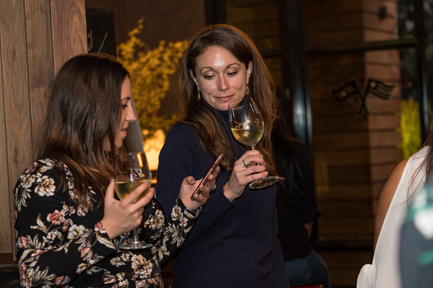 Two women hold wine glasses and look down at a phone against a wooden wall at a bar, one in a floral top and one in a navy sweater.