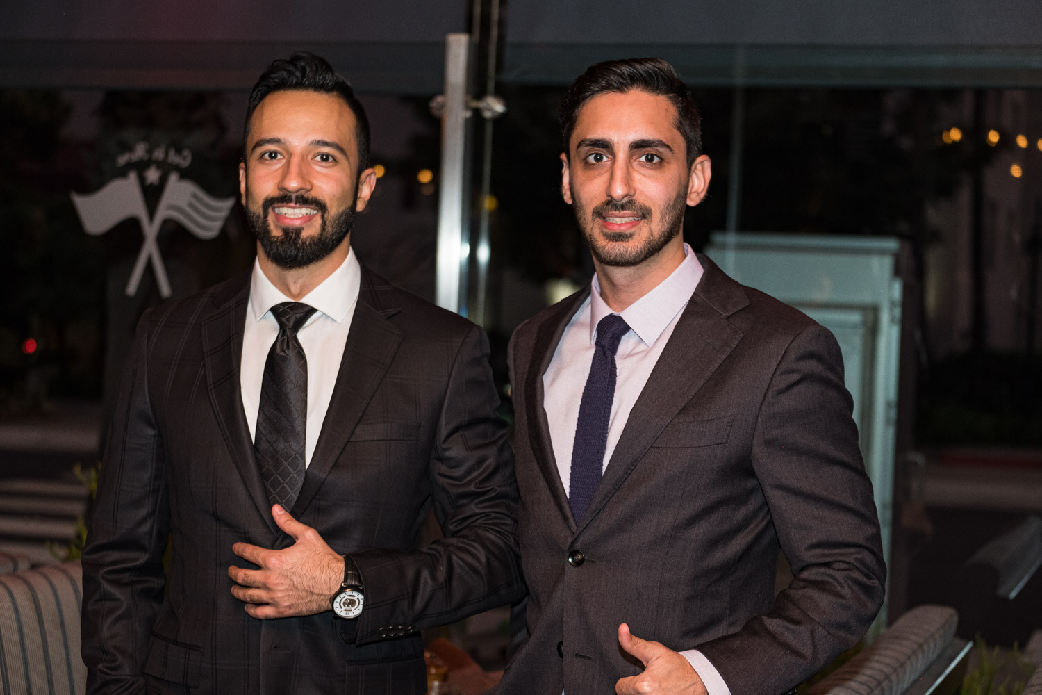 Two smiling men in dark suits and ties stand side by side in front of a window at night.