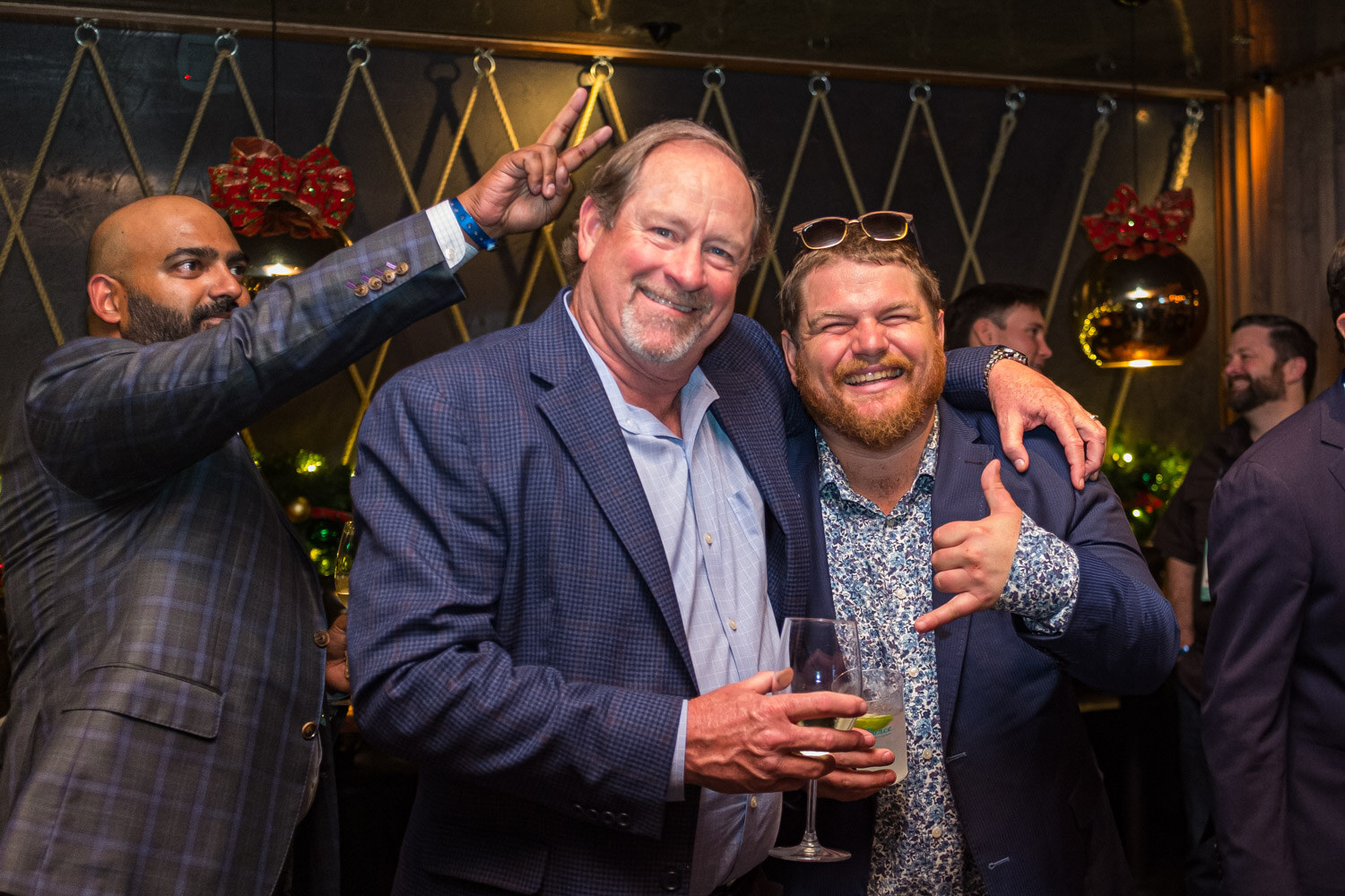 Three men in blazers pose arm in arm holding drinks in front of a dark wall with rope lattice and red holiday bows, one making a peace sign behind another.