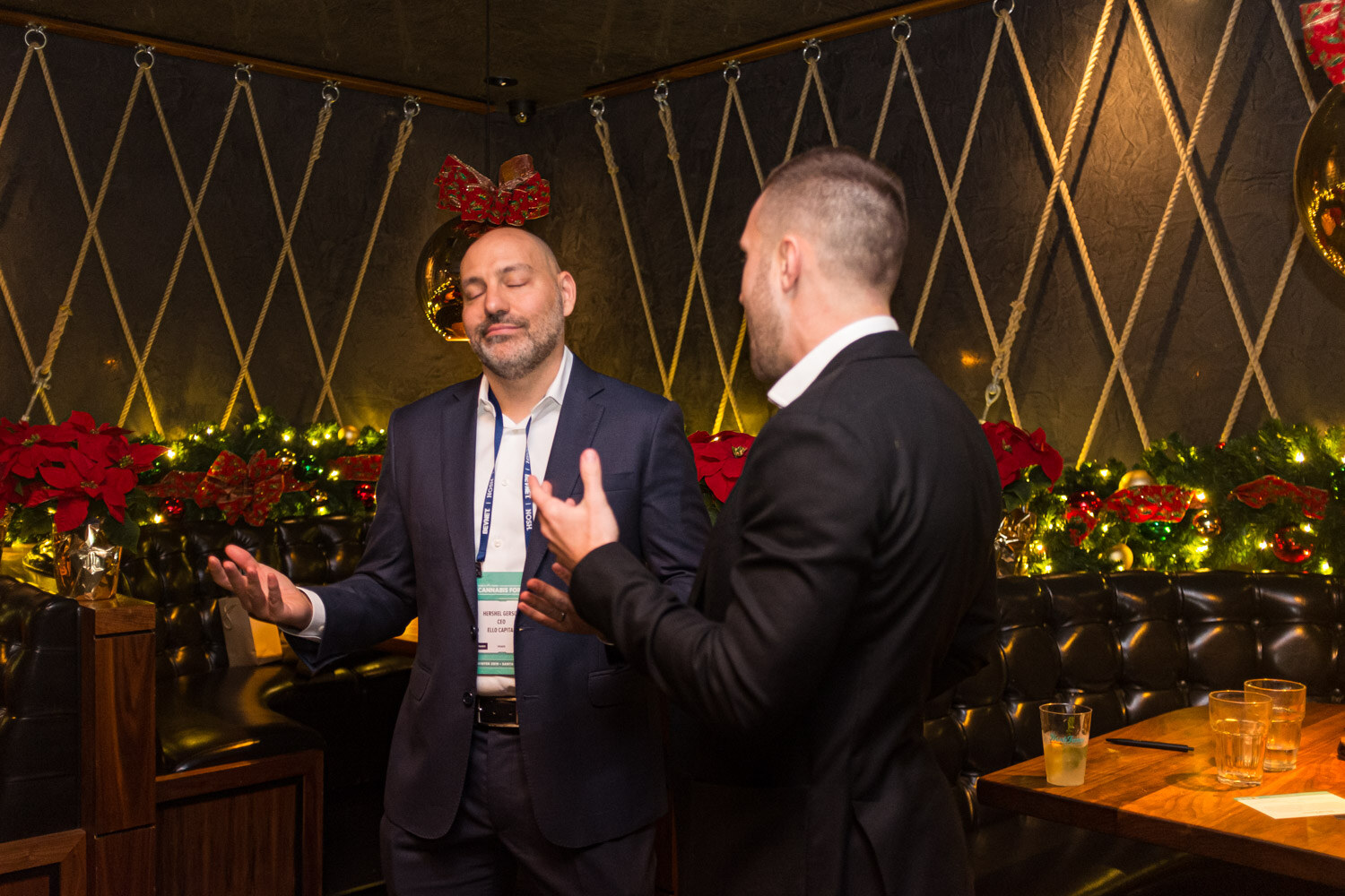 Two men in dark suits talk with open hands, one wearing a conference lanyard, in front of a leather booth with red poinsettias and green garlands.