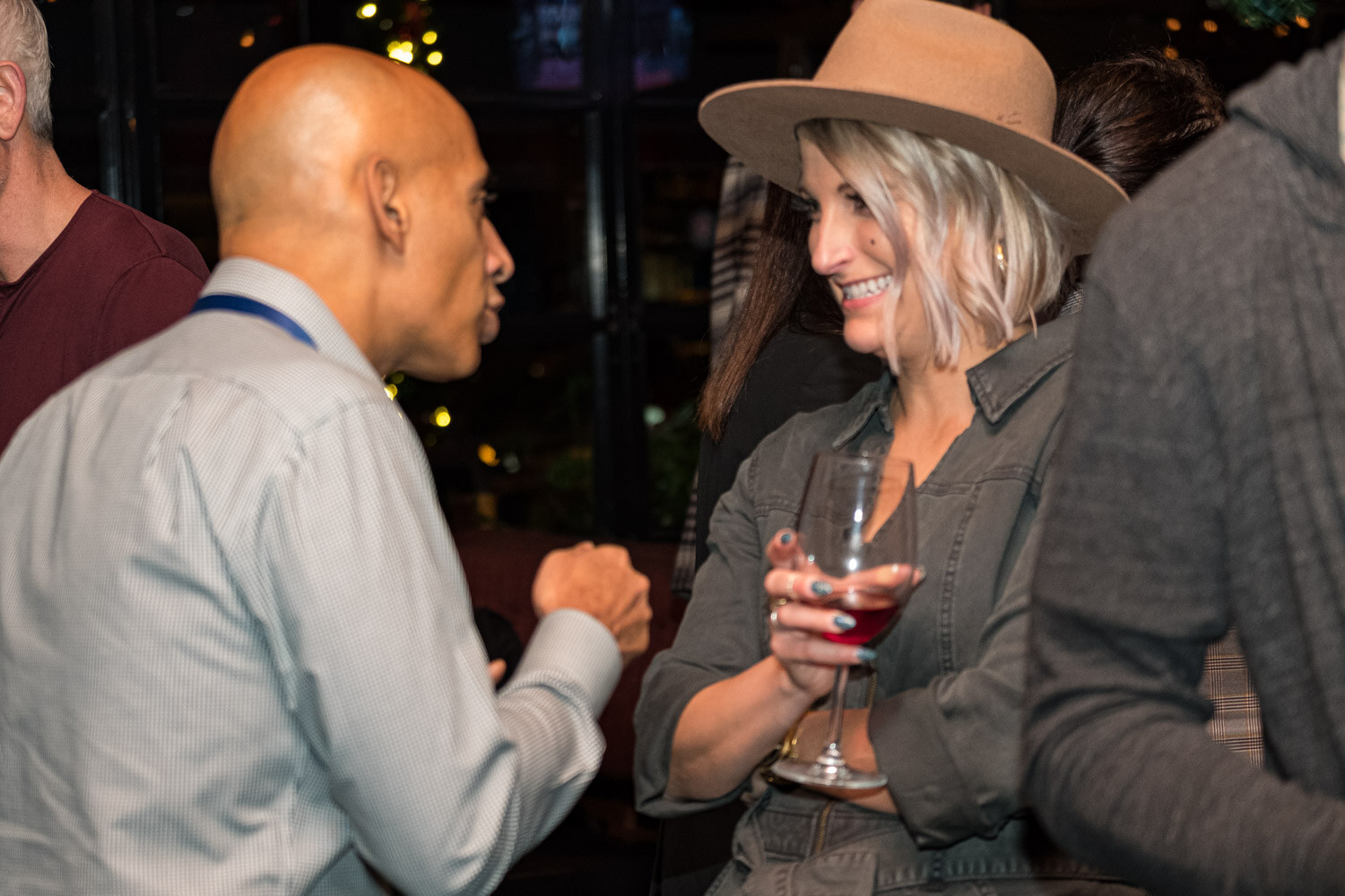 A bald man in a light gray shirt talks with a smiling woman in a tan felt hat holding a glass of red wine, with other guests around them.