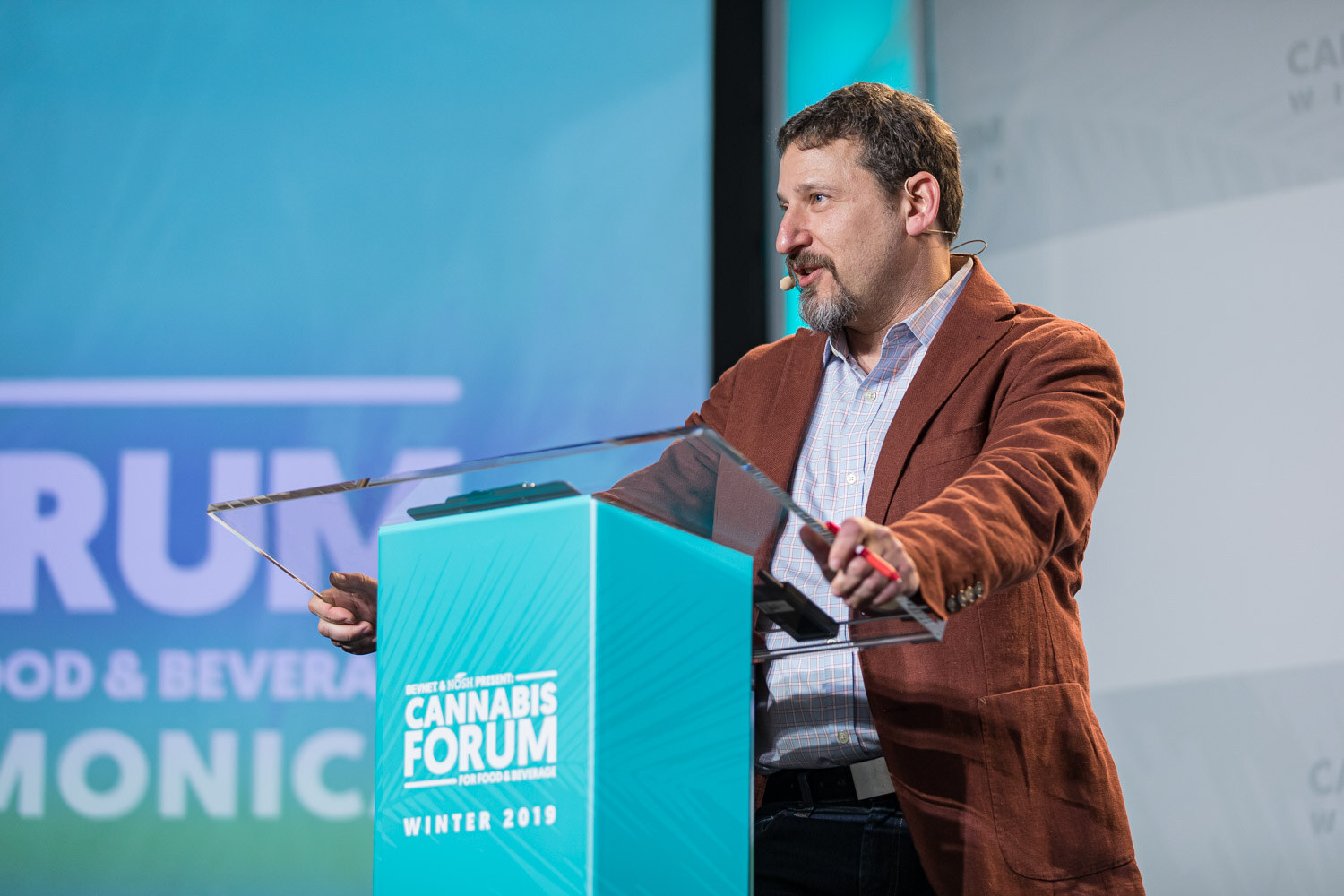 A bearded man in a rust-colored corduroy blazer speaks at a teal Cannabis Forum podium in front of a teal screen.