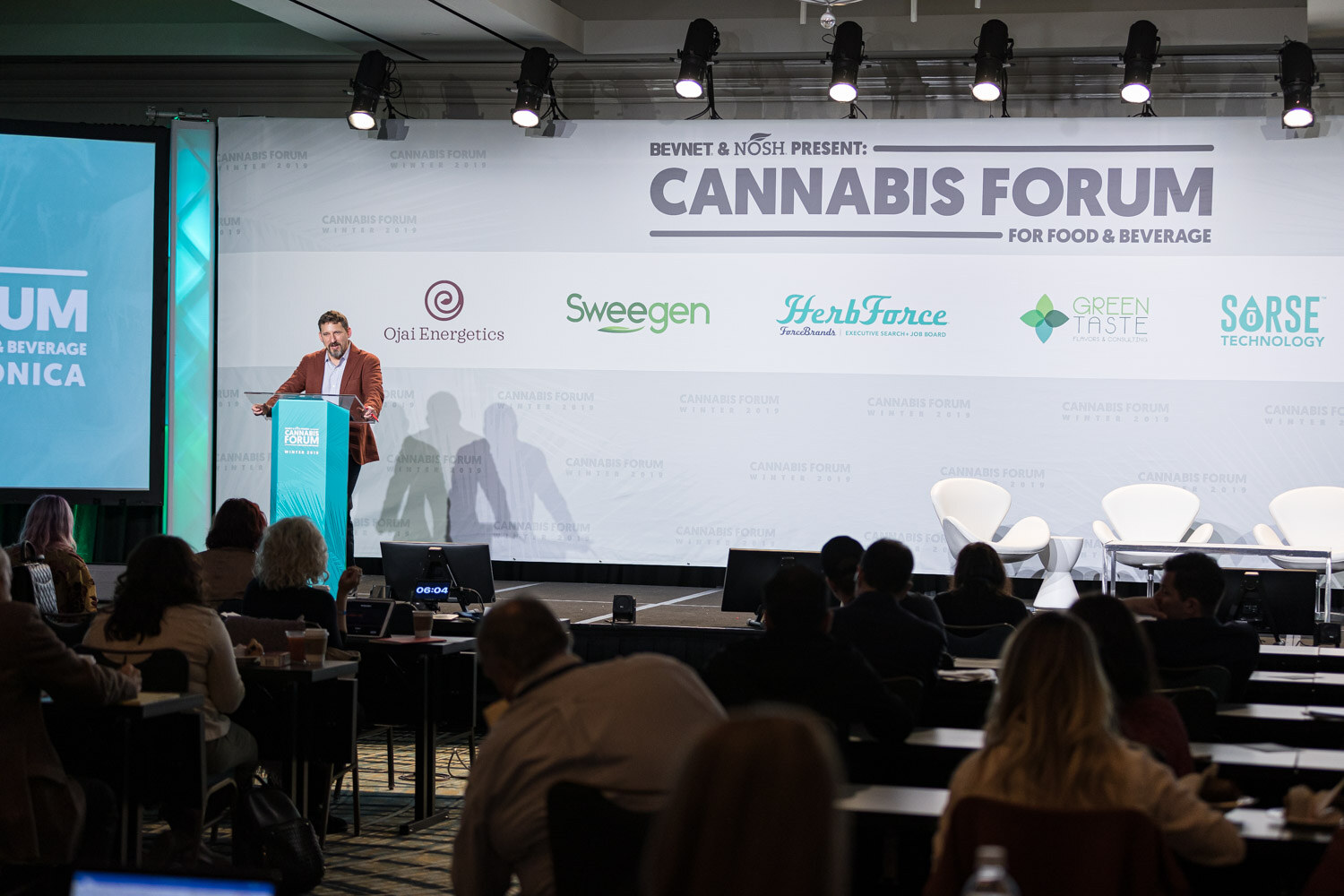 A wide view of the ballroom shows a speaker at the podium on stage in front of a Cannabis Forum for Food & Beverage backdrop with Ojai Energetics, Sweegen, HerbForce, Green Taste and Sorse logos, and seated audience in the foreground.
