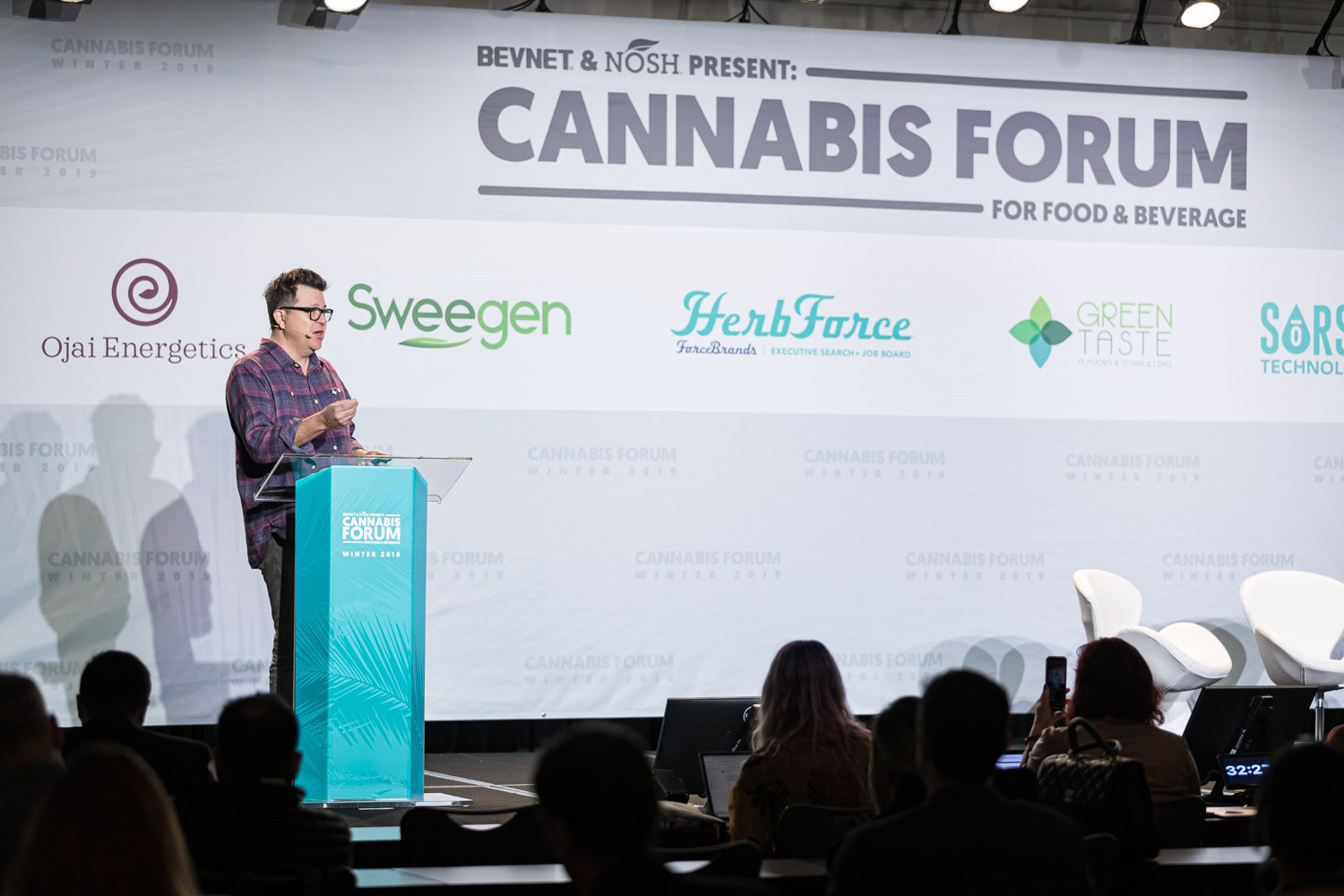 A man in a plaid shirt stands at a teal podium on a stage in front of a banner reading BevNET & Nosh Present Cannabis Forum for Food & Beverage, with silhouetted attendees in the foreground.