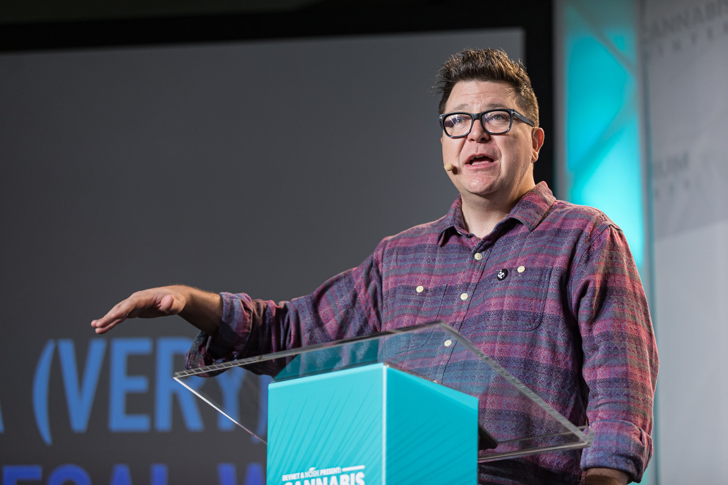A man in glasses and a plaid shirt reaches one arm out over a glass podium, with blue lettering from a projected slide behind him.