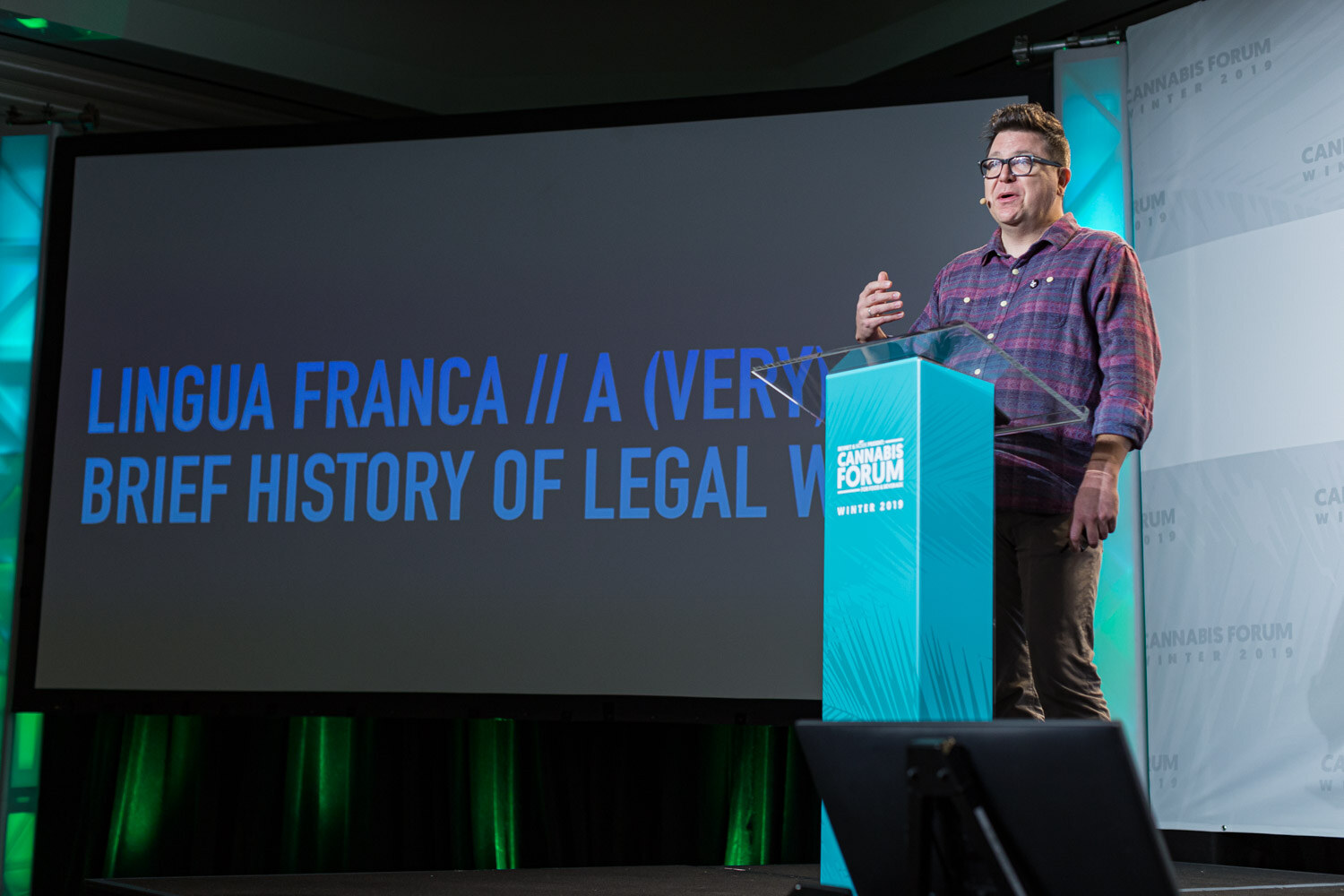A man in a plaid shirt speaks at a teal podium beside a projection reading Lingua Franca // A (Very) Brief History of Legal Weed.