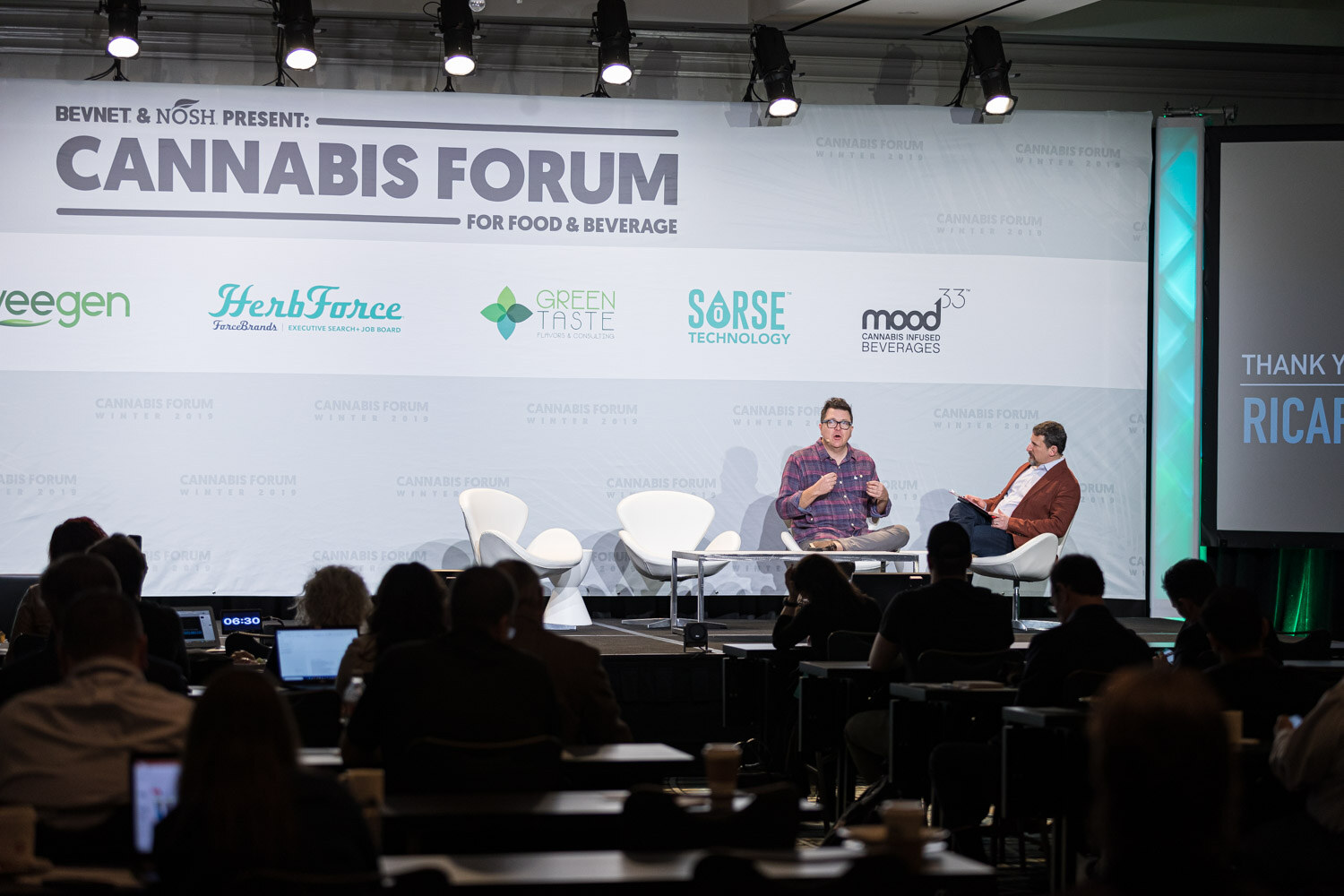 Two men sit in white lounge chairs on a stage in front of a Cannabis Forum backdrop, with silhouetted attendees and laptops in the foreground.