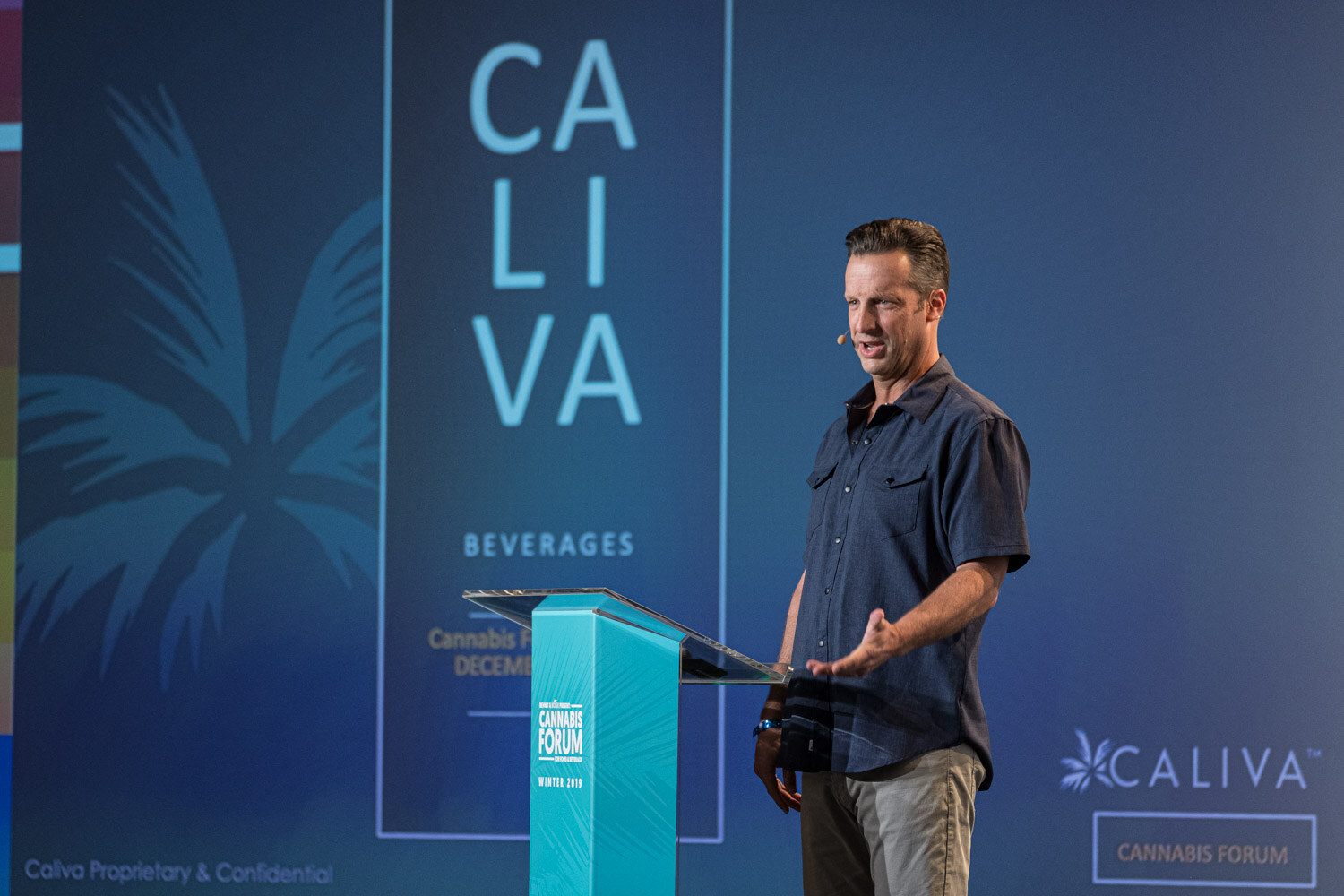 A man in a dark short-sleeved shirt stands at a teal podium beside a blue slide with the Caliva Beverages logo and a palm frond design.