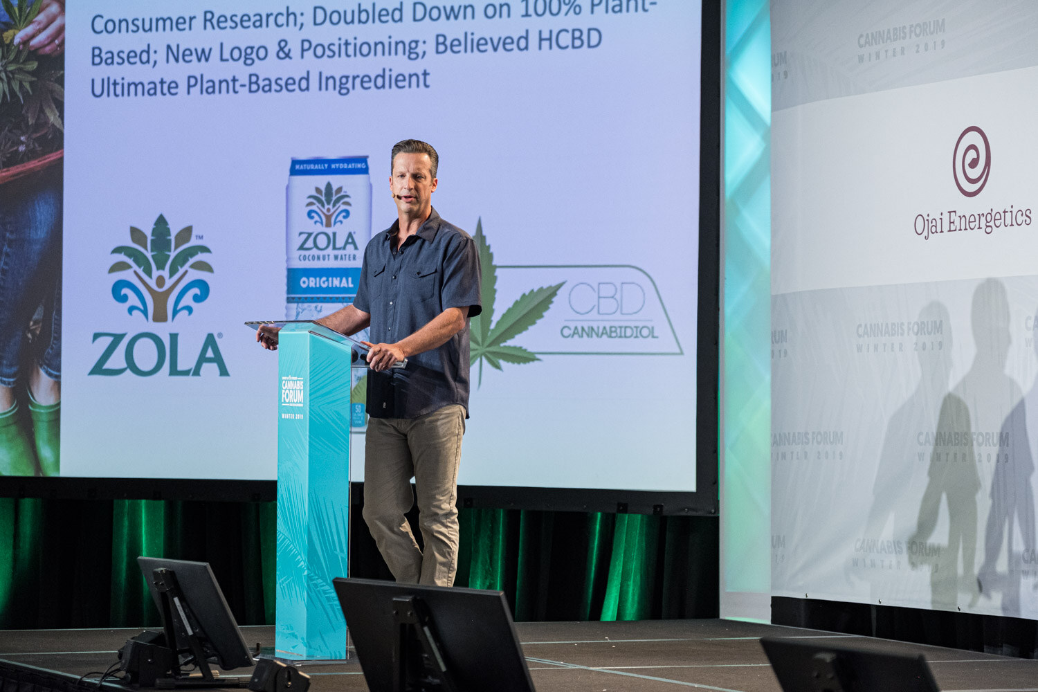 A man in a dark short-sleeved shirt stands at a podium in front of a large slide with the Zola logo, a Zola coconut water carton and a CBD cannabidiol mark.